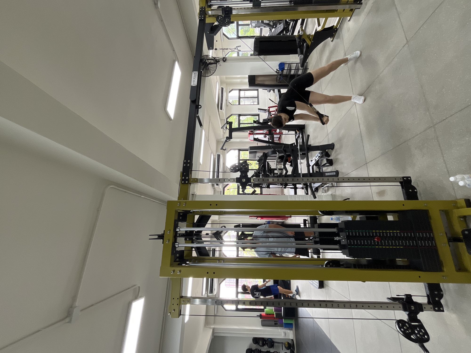 The image shows an indoor gym with modern exercise equipment. In the foreground, a woman in black athletic wear is performing a cable crossover exercise, holding a handle connected to a weight machine. To the left, two men are standing near other weight machines. One man in a black t-shirt and blue shorts is looking away, while another man in a grey t-shirt and black shorts is facing a weight machine. The gym features large windows with a view of buildings and trees, suggesting an urban setting. The lighting is bright, with overhead fluorescent lights and natural light filtering through the windows. The floor is a light grey tiled surface. The presence of gym equipment and the overall ambiance suggest a fitness facility in Chiang Mai City Municipality, Thailand.
