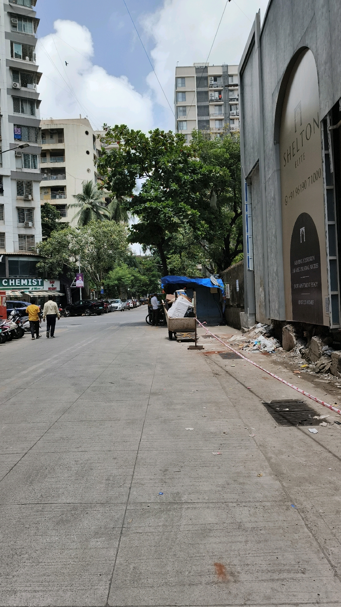 A street scene in Mumbai, India, on a cloudy day. A broad, grey concrete road runs from the foreground into the distance, with white road markings. On the left, a white multi-story apartment building is visible, with a sign for "CHEMIST" in green and white text. To the right of the chemist, a smaller shop front is visible with a poster featuring a man's face. Further down the street, parked motorcycles line the left side of the road, and several cars are parked on the right.
Two men are walking away from the camera on the left side of the road. One is wearing a yellow shirt and dark pants, the other a pale yellow shirt and dark pants.
On the right side of the road, a building with a grey facade and large arched windows is prominent. A sign on this building reads "SHELTON ELITE" and includes a phone number. Adjacent to this building, a makeshift shelter covered with a blue tarp is set up, with debris and discarded items piled nearby. A trolley filled with what appears to be trash or construction materials is parked next to the shelter. Red and white caution tape is stretched along the side of the road near the shelter and the building. Tall green trees with lush foliage are visible throughout the scene, interspersed between the buildings. The sky is overcast with scattered clouds.