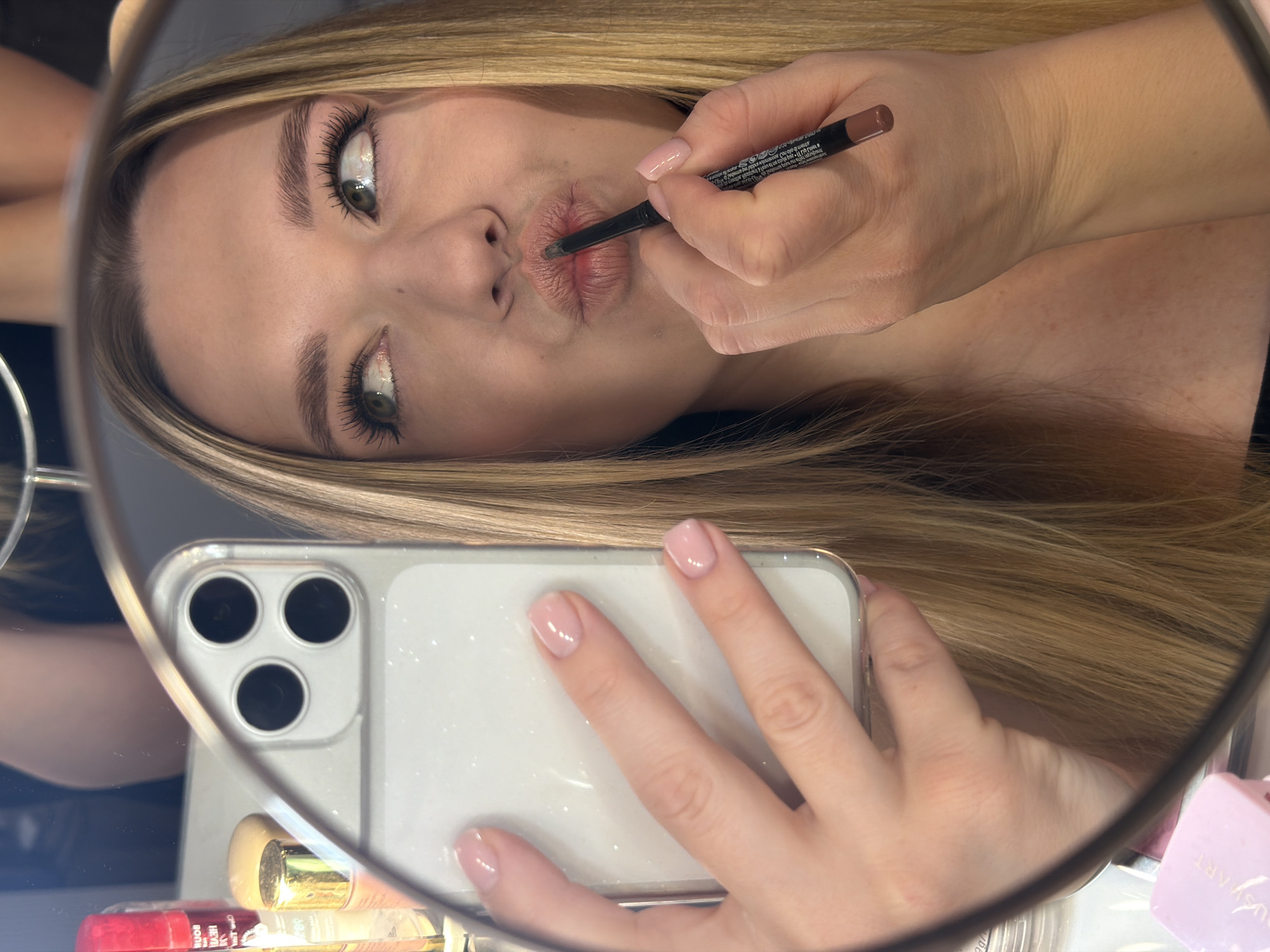 The image depicts a young woman reflected in a round mirror, taking a selfie with a white iPhone. She is applying lip liner. The location is not determinable from the image, and it is not possible to confirm it is Dreiliņi, Latvia.