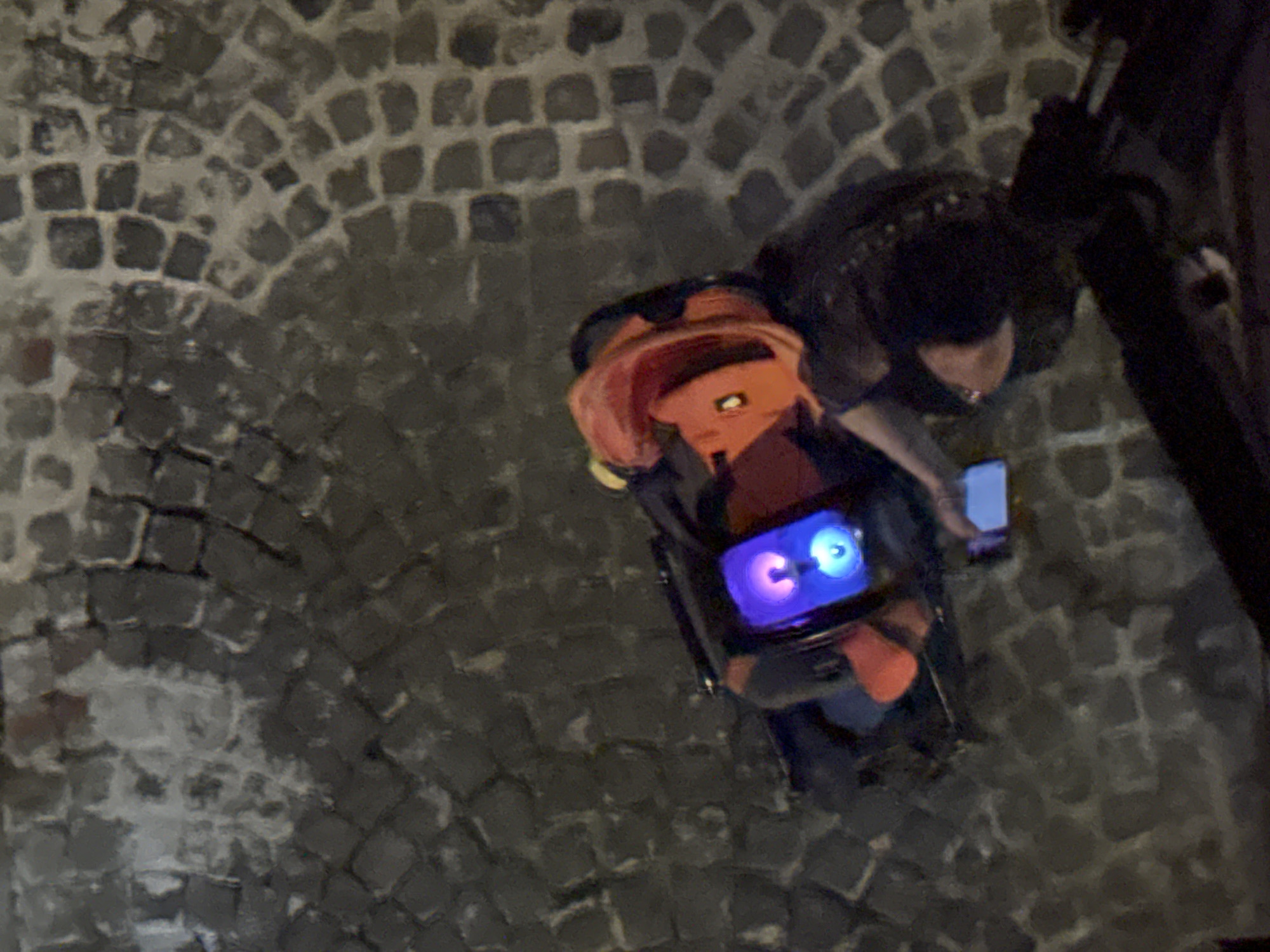 Observed from an elevated perspective on what appears to be a cobblestone street, potentially in Castelnuovo di Porto, Italy, the scene depicts an individual standing beside a brightly lit, compact object. The individual is partially visible, appearing to interact with a mobile phone in their hand. The brightly lit object is equipped with a screen displaying glowing, cartoonish eye-like features. The area surrounding the individual and the object is dimly lit, with the cobblestone pavement forming a significant portion of the visual background.