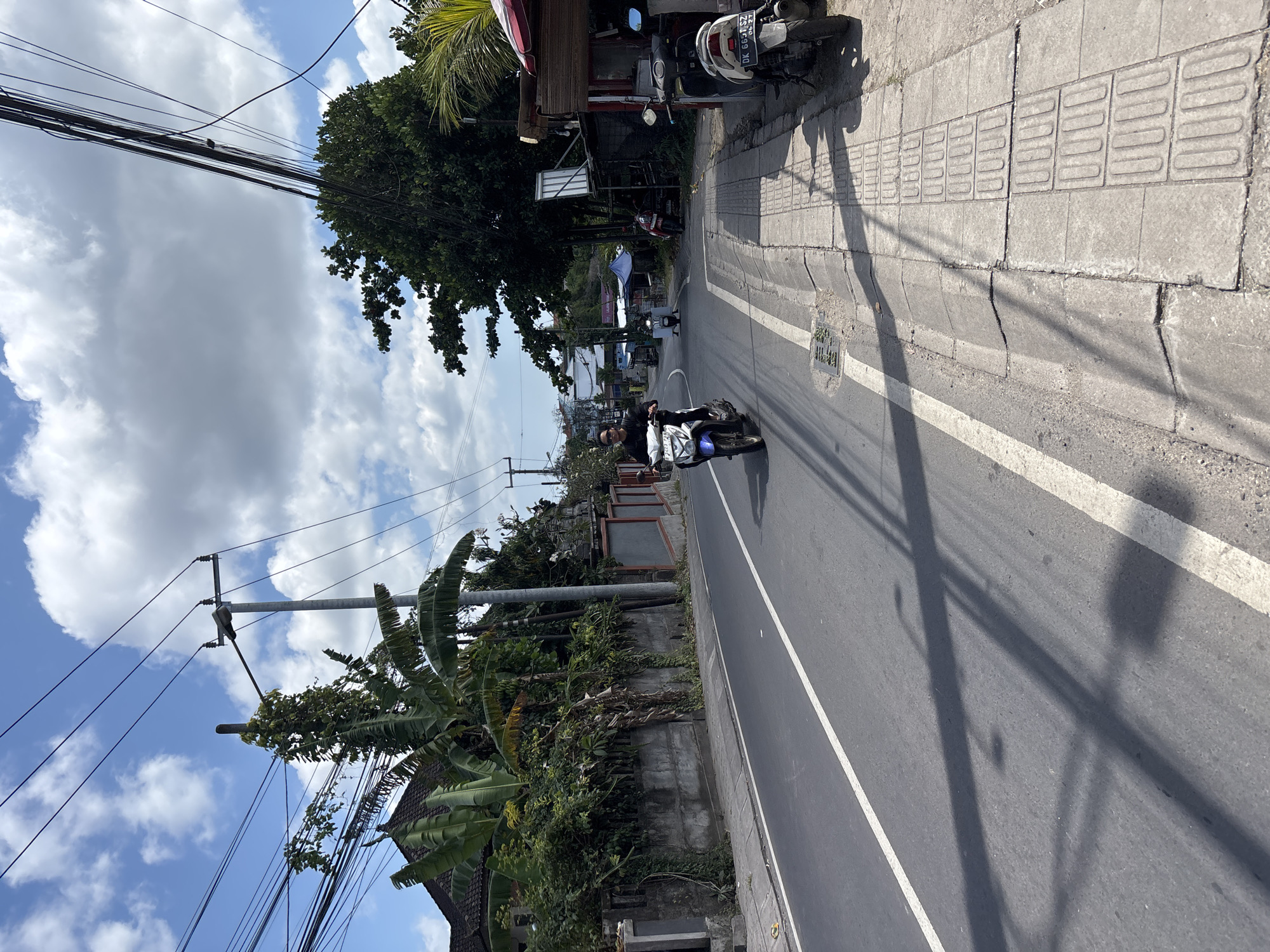 An outdoor, vertical street scene depicts a road and sidewalk bordered by lush tropical vegetation and buildings under a cloudy but bright sky. Long shadows are cast across the road and sidewalk, suggesting it is daytime, possibly morning or late afternoon.
In the mid-ground, a person wearing a dark shirt and sunglasses is riding a blue and white motorcycle away from the viewer on the asphalt road, which features a single solid white line. On the right side of the road, a concrete sidewalk with distinctive rectangular tactile paving is visible. Several parked motorcycles and scooters are present along the sidewalk further down the street, one of which prominently displays an Indonesian license plate "DK 6641 SZ" with "08.27" below it, indicating an expiry date.
The left side of the scene is dominated by a variety of green trees and plants, including banana trees and dense foliage, which partially obscure the concrete walls and roofs of buildings. Utility poles with numerous overhead power lines stretch across the frame, silhouetted against the sky. The overall setting captures a typical street environment in Kuta Selatan, Indonesia.