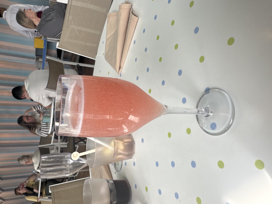 The scene depicts the interior of a casual dining establishment or cafe in Puerto del Rosario, Spain. In the immediate foreground, a clear, tall-stemmed glass containing a sparkling, light pink beverage with visible bubbles stands centered on a white tabletop. The tabletop features a scattered pattern of light blue and light green polka dots. To the left of this glass, a shorter, dark plastic cup holds a dark liquid, and a frosted plastic tumbler contains a light, clear liquid with a white plastic stirrer. A folded beige fabric napkin rests to the right of the central glass.
In the mid-ground and background, multiple individuals are seated at tables. On the left, a woman with dark hair wears a dark top and appears to be drinking from a glass. Behind her, another individual with light hair is partially visible. Further right, a woman with dark hair and glasses, wearing a horizontal striped shirt, is seated and looking down at a mobile phone. Across from her, a man with short dark hair, wearing a vertically striped shirt, is also seated and appears to be looking down. On the far right, a woman with light hair and glasses, dressed in a grey top, is seated and looking to her right. Behind her, a man in a white collared shirt, potentially a staff member, is standing. The background wall or partition displays vertical stripes in alternating shades of light blue, pinkish-beige, and white. The overall environment suggests a relaxed atmosphere typical of a cafe or restaurant.