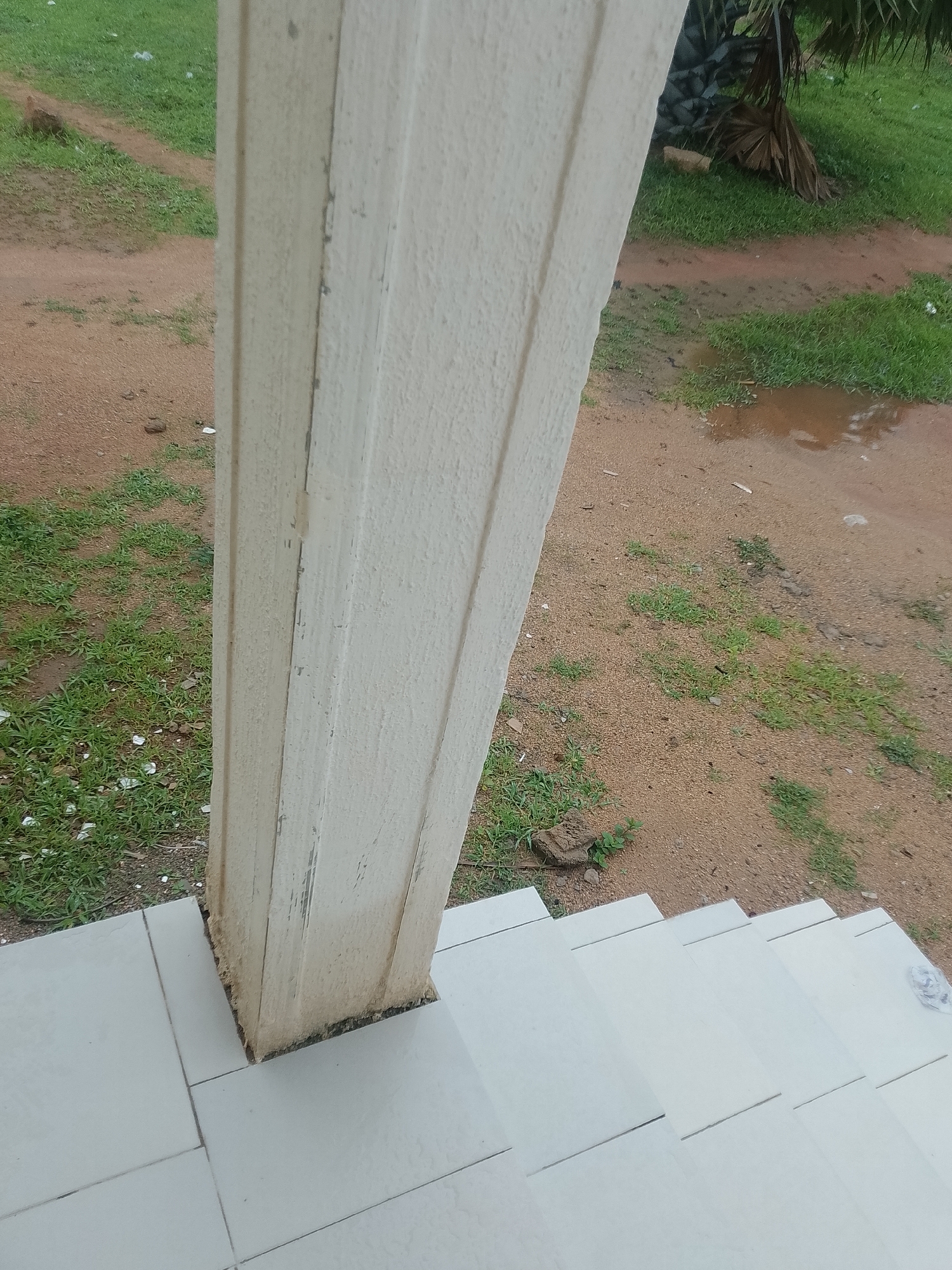 A close-up, oblique, eye-level shot focuses on a cream-colored, textured rectangular column situated on a set of pale gray tiled stairs. The column has a rough surface with some scuff marks and dirt at its base. The stairs appear to be outdoors, with a dirt path and patchy green grass visible behind the column. A large puddle of muddy water is present in the background, along with a palm tree. The ground is a mix of reddish-brown dirt and scattered green grass. The overall scene suggests an outdoor environment, possibly in a tropical or subtropical region.