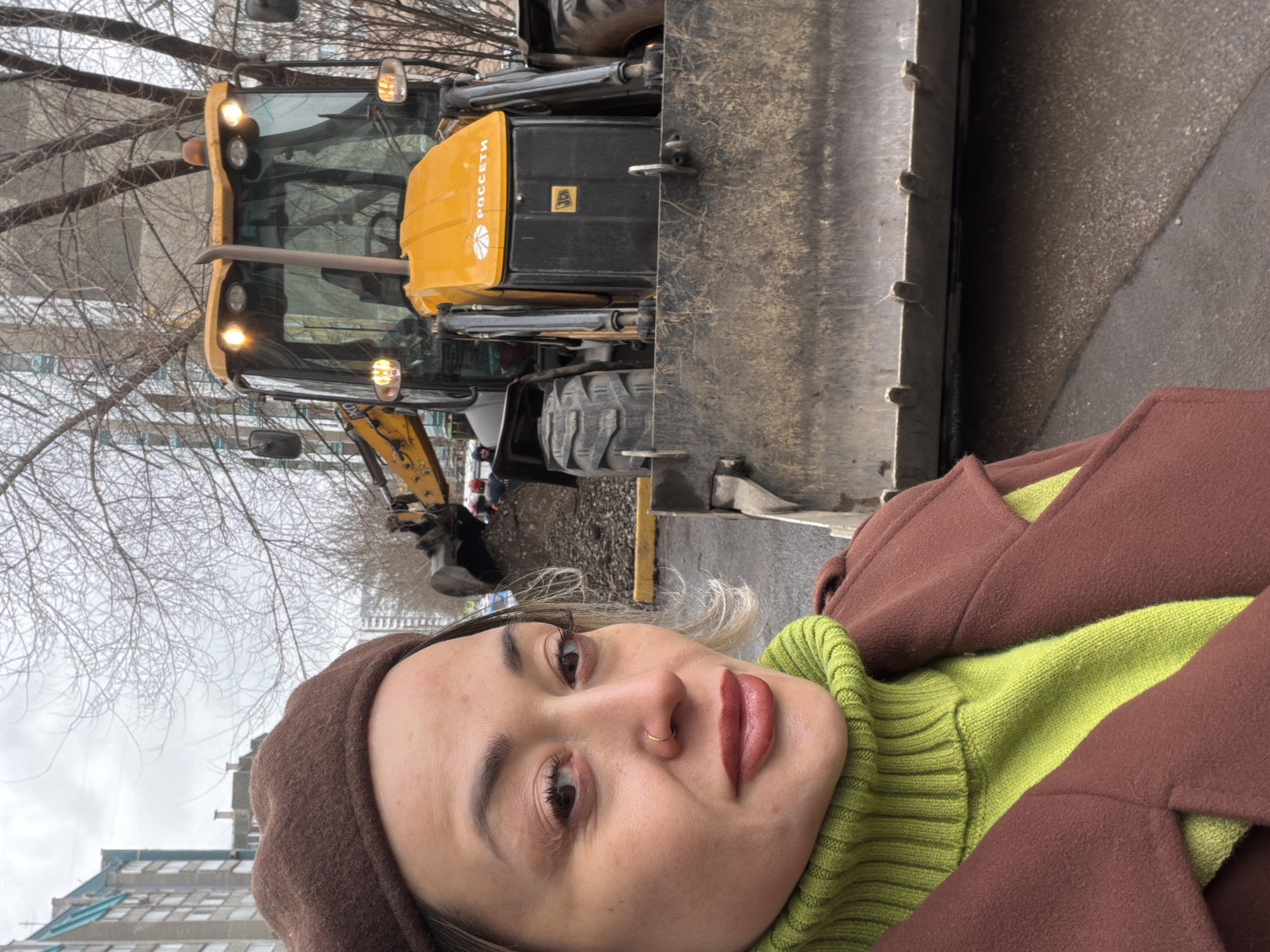 The photograph depicts a woman posing in front of a yellow and black backhoe in an urban setting. The woman is wearing a brown beret, a green sweater, and a brown coat. The backhoe has "POCCETИ" (Rosseti) in Cyrillic script on its side, indicating a Russian origin. Bare trees and a building with light blue accents are visible in the background. This location can be identified as Krasnoyarsk, Russia due to the presence of "POCCETИ" vehicles that work in the area. A small construction site is visible in the background.