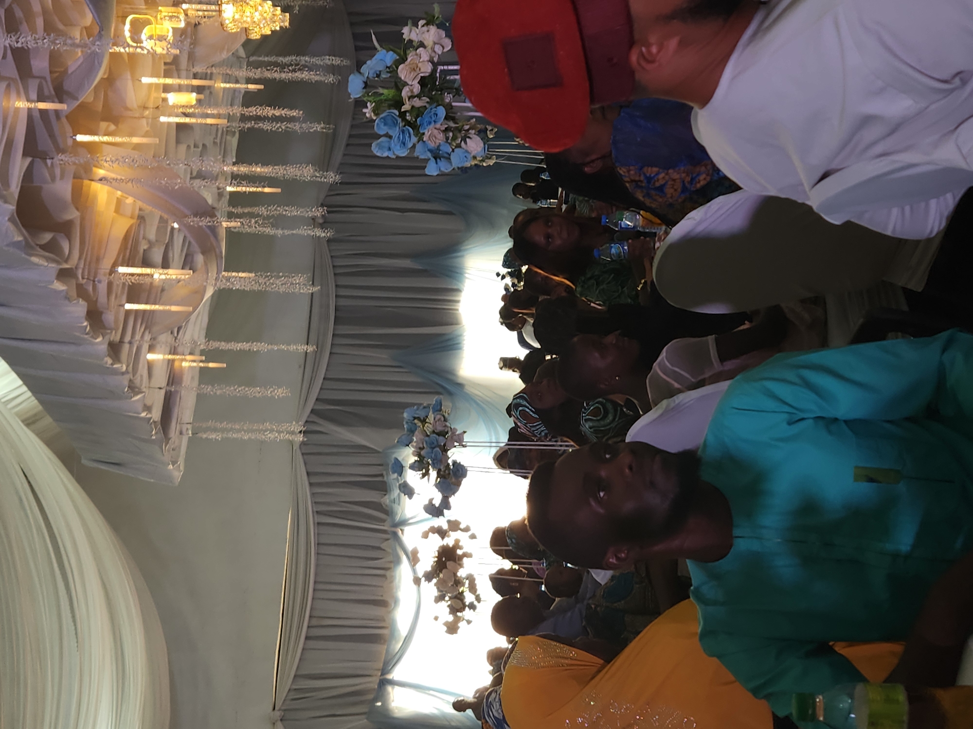The image captures an indoor scene in Jos, Nigeria, filled with people attending an event. Individuals of varying ages and genders are seated, engaging in conversation and interactions. The setting appears to be decorated for a special occasion, evident by the adorned ceiling with draped white fabric, chandeliers, and floral arrangements in blue and white hues. Some individuals are wearing traditional Nigerian attire, including a man in a red cap. A person in a teal shirt is in the foreground of the image. The image suggests a celebratory atmosphere.