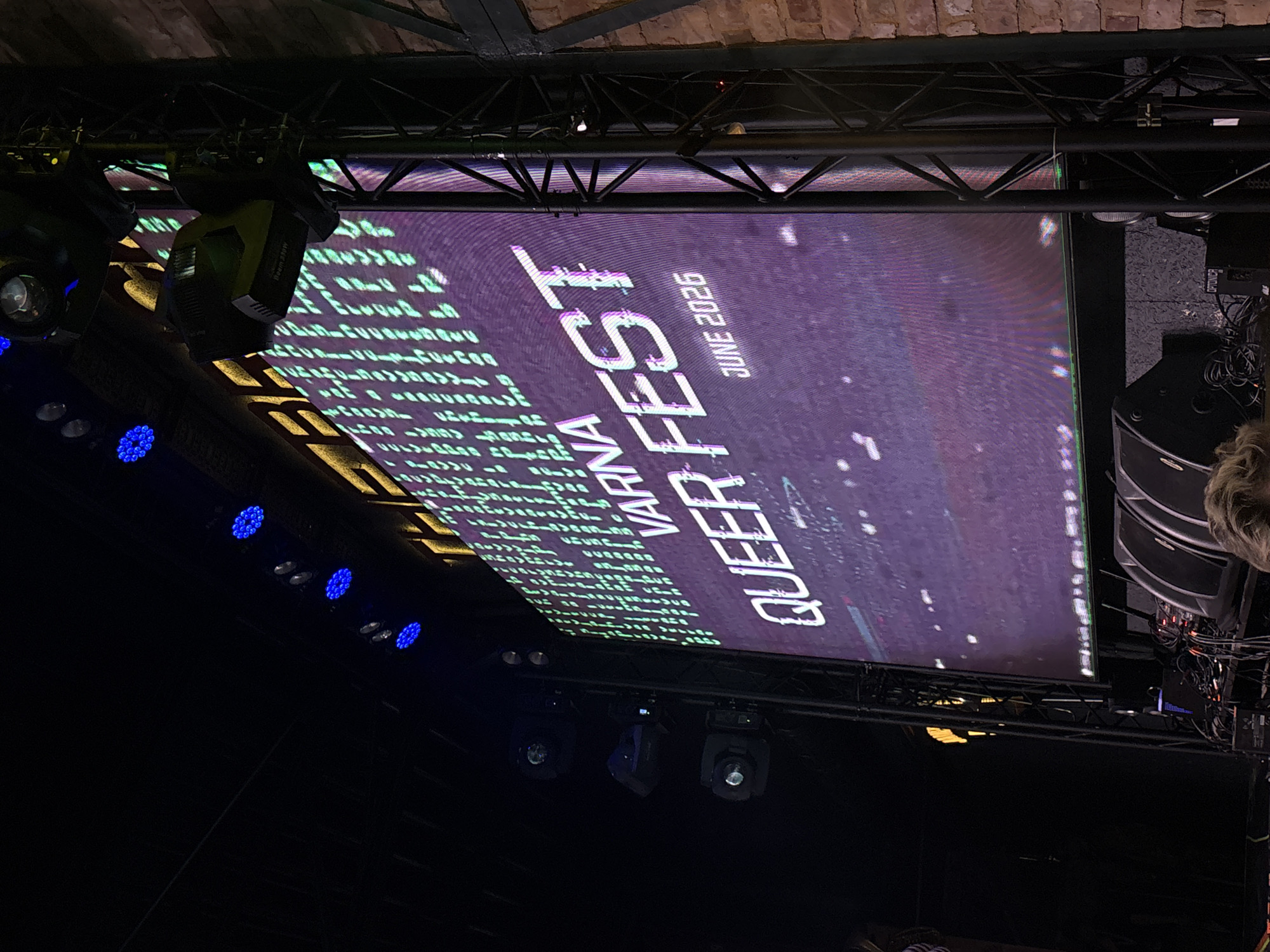The image shows a large LED screen displaying the text "QUEER FEST VARNA JUNE 2026" in bold white font against a purple, speckled background. To the left of the main text, there is a column of green, pixelated text that appears to be code or a similar digital pattern.
The screen is positioned above a stage or performance area, with stage lighting fixtures visible below and to the left. Several blue stage lights are illuminating the lower left portion of the image. Metal trussing structures are visible above the screen, supporting the lighting and possibly other stage equipment.
The setting appears to be an indoor venue, possibly a club, theater, or conference hall, given the presence of stage lighting, sound equipment (speakers are visible on the right), and a brick wall in the background. The lighting is dim, characteristic of an event at night or an indoor venue. There are no people clearly visible in the main focus of the image, although a person's hair is visible on the far right, suggesting an audience member or technician. The overall mood is celebratory and modern, indicated by the bright screen and stage lighting. The text provides specific information about the event, its name, location, and date.