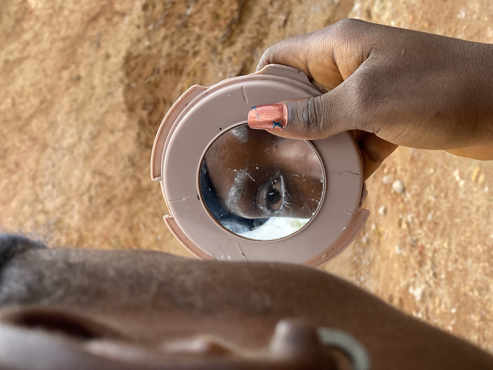 A close-up, over-the-shoulder shot shows a person's left hand holding a round, light-pink compact mirror. The mirror reflects the right eye and eyebrow of the person, who appears to be looking into it. The person's skin is dark, and the eye in the reflection is also dark. The eyebrow is well-defined and slightly arched. The person's fingernail on their right index finger, which is visible in the frame and pointing towards the mirror, is painted a coral-orange color with green metallic accents.
The background consists of a dry, reddish-brown, rocky terrain, indicative of a semi-arid or desert environment. The ground is rough and uneven, with some scattered pebbles and debris. The lighting suggests it is daytime, with a soft, diffused light source casting subtle shadows. The overall impression is one of a candid moment captured outdoors, possibly in a rural or less developed area. The specific location within Bar Jirgi Summa, Nigeria, is not discernible from the visual information alone.