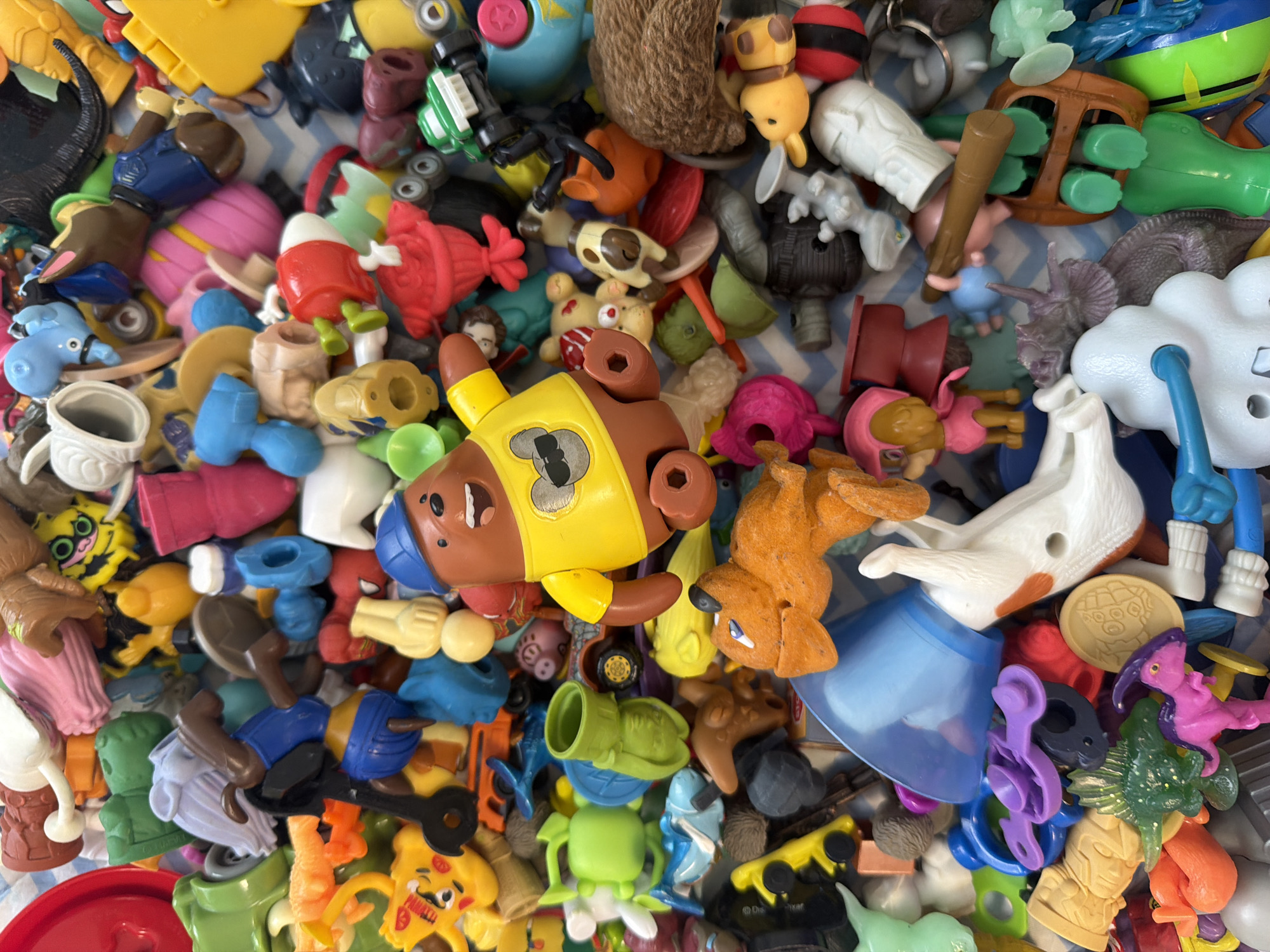 The image depicts a jumbled collection of plastic toys and toy parts. The toys vary in color, size, and form, including figures of animals, characters, and abstract shapes. There are identifiable elements such as a yellow bear figurine wearing a vest, a white horse-like figure, and various components suggesting a diverse range of toy sets. The background is a blue and white patterned surface. There is no discernible evidence to connect this collection to the city of Votkinsk, Russia.