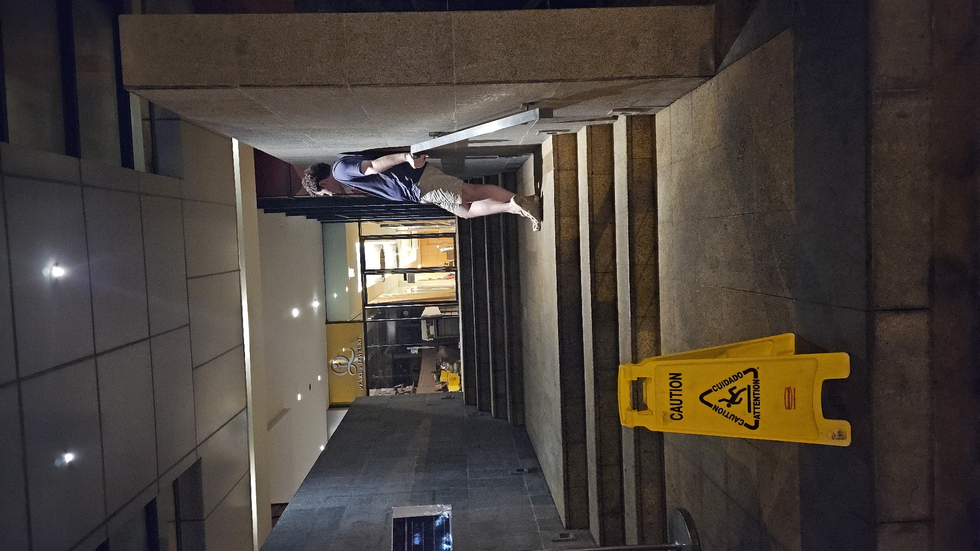 A medium shot of a man descending a set of stairs in Nha Trang, Vietnam. The man is holding onto the railing with both hands. He is wearing a dark blue shirt and khaki shorts. His dark blond hair is closely cropped. There is a yellow caution sign at the bottom of the stairs. The sign reads "CAUTION" on both sides, and also has the Spanish word "Cuidado". The stairs lead down to a building with the words "PEARLA JEWELRY" prominently displayed. The building's windows are brightly lit. The stairs are made of gray stone. The environment is urban, and the time of day appears to be evening or night.