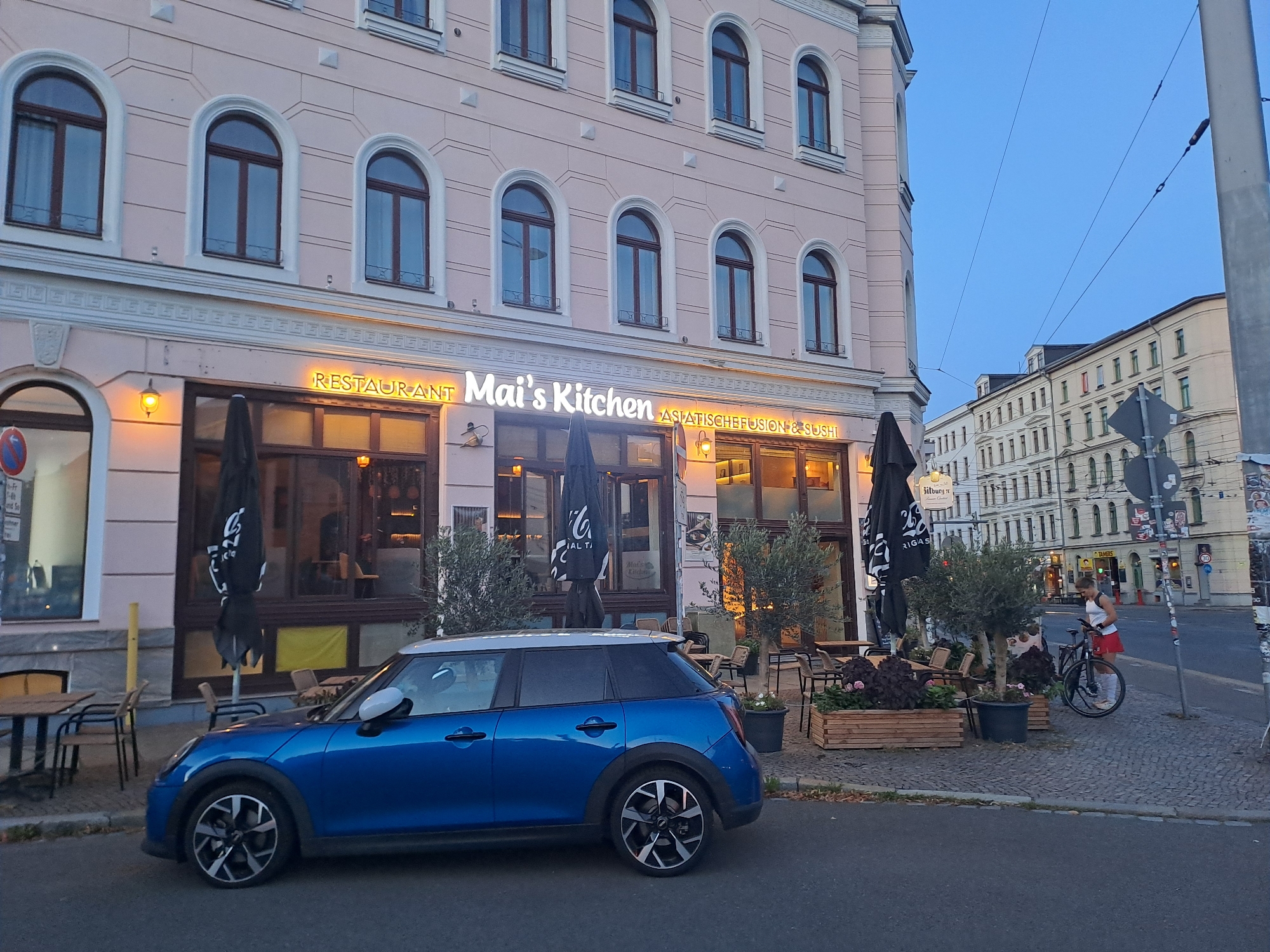 The image depicts a street scene in Leipzig, Germany, at dusk. The foreground is dominated by a blue Mini Cooper parked on the street. To the right of the car is a sidewalk with several potted plants and outdoor seating for a restaurant. The restaurant, "Mai's Kitchen," is visible with its illuminated signage. The building behind the restaurant has a pale pink facade with arched windows. Further down the street, other buildings are visible, as well as a person standing with a bicycle. Overhead power lines suggest the presence of public transportation. The sky is a clear, pale blue.