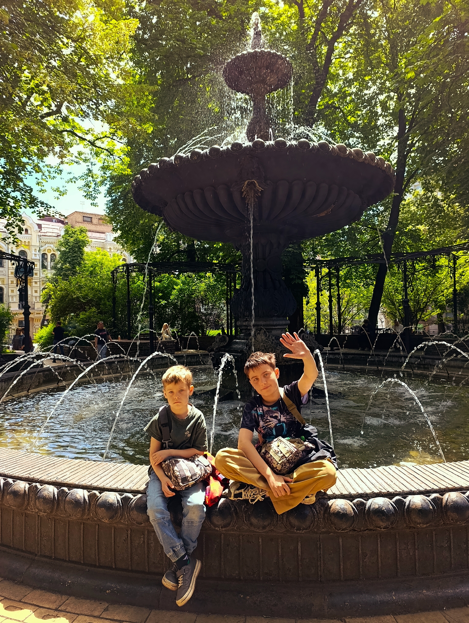 Two boys are sitting on the edge of a tiered fountain in a city park. The boy on the left, wearing a green t-shirt, blue jeans, and gray sneakers, holds a bag on his lap. The boy on the right, wearing a dark t-shirt and ochre pants, is waving. Both boys have bags. The fountain is surrounded by trees, and buildings can be seen in the background. The location is Kyiv, Ukraine.