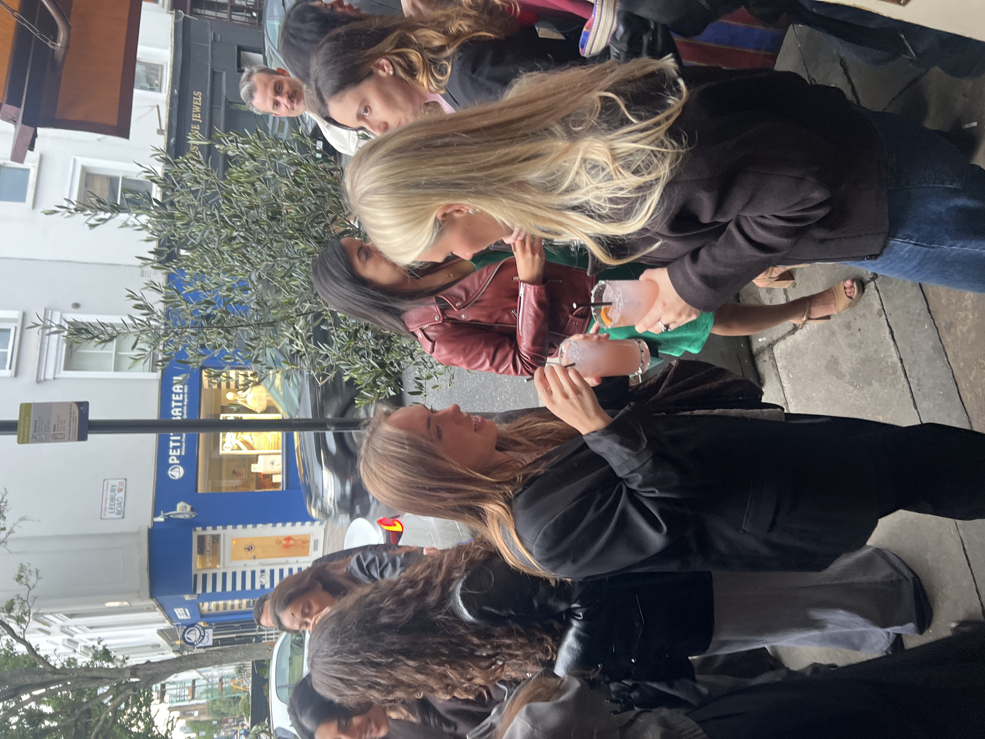 A street scene in London, United Kingdom shows a group of approximately 10 people standing on a sidewalk outside a building. The people appear to be of mixed ages and ethnicities. They are generally dressed in casual clothing, including jackets, jeans, and skirts. Several individuals are holding drinks in glasses.
The buildings in the background feature shops and businesses, identifiable by signage. One building displays the sign "PETIT BATEAU." There is a street sign that reads "LEDBURY ROAD W11."
A tree is visible between the people and the buildings, adding a touch of greenery to the urban scene.
The time of day appears to be late afternoon or early evening, judging by the soft lighting. The sidewalk is paved, and the area appears clean and well-maintained.
The group of people appear to be engaged in a social gathering, possibly enjoying drinks or conversation together.