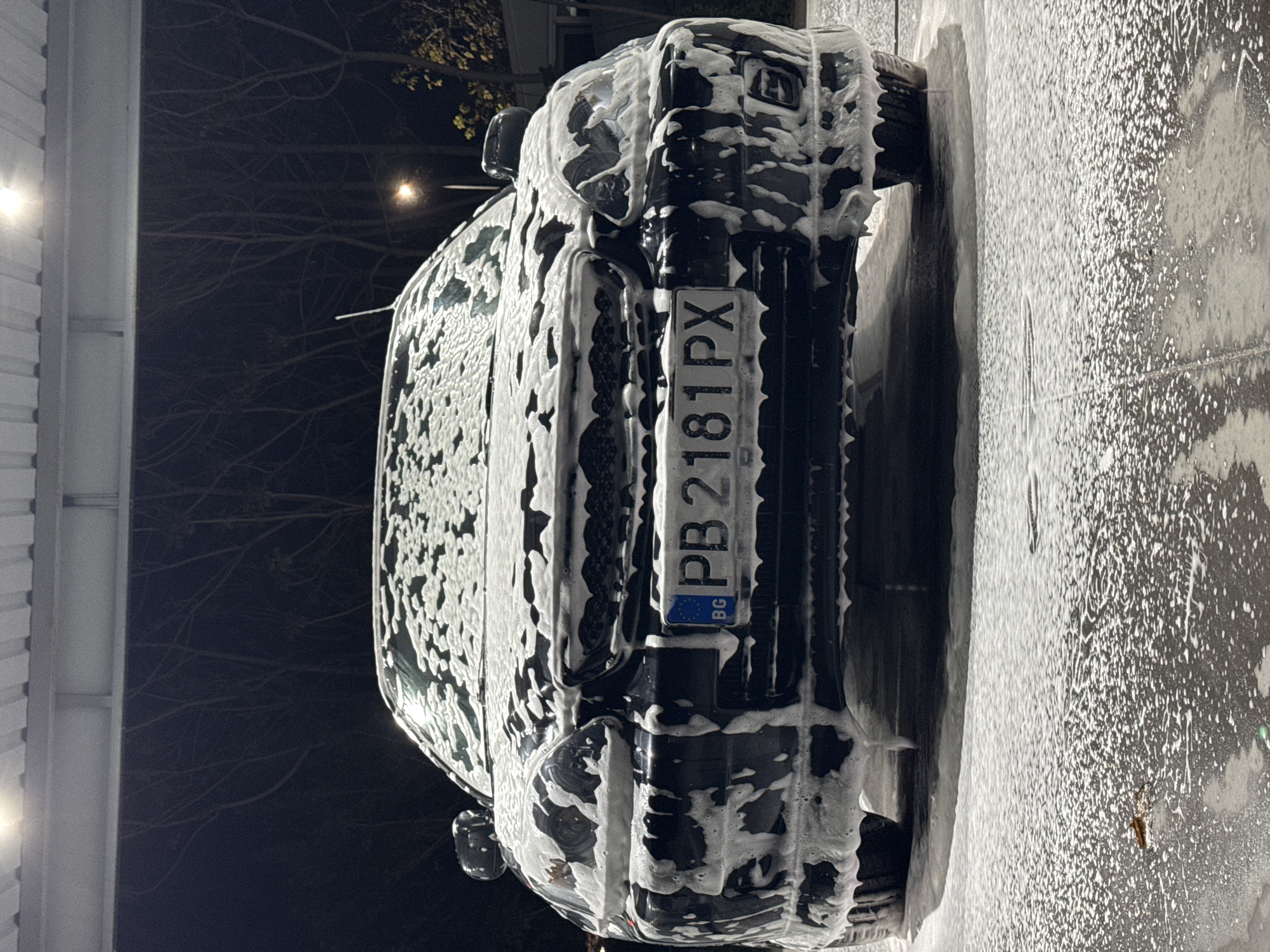 The image shows the rear of a black car covered in white soap suds, indicating it is undergoing a car wash. The license plate reads "PB 2181 PX," and a small "BG" emblem is visible, confirming it is a Bulgarian vehicle. The surrounding environment suggests an outdoor setting at night, with a building and trees visible in the background, and the presence of artificial lighting. The location in Plovdiv, Bulgaria, can be inferred from the "PB" designation on the license plate, which corresponds to the Plovdiv region.