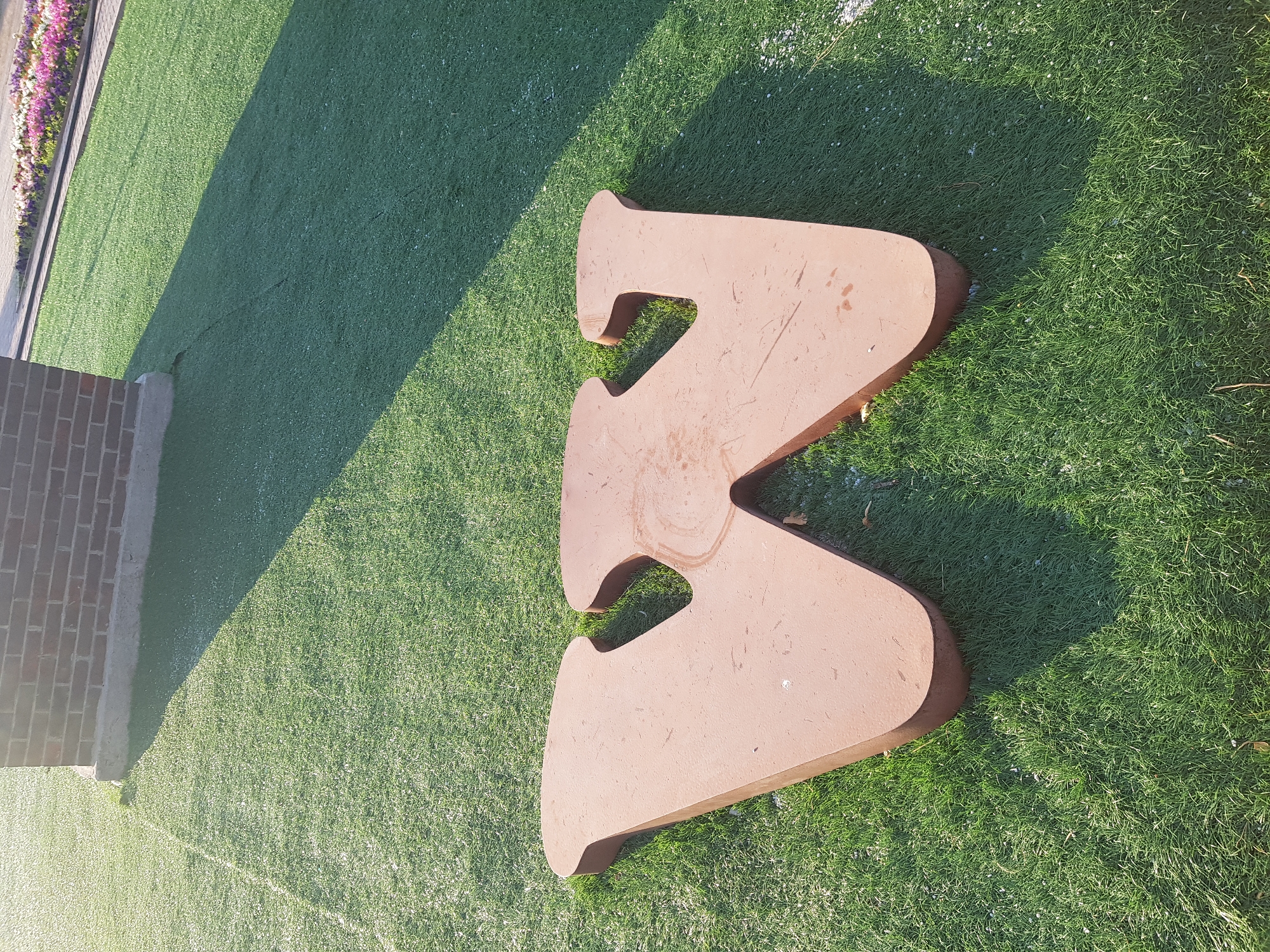 A large, three-dimensional metal letter "W" is situated on a patch of artificial grass. The letter is a reddish-brown color and appears to be made of corten steel or a similar material that has developed a patina. It has a rounded, slightly weathered appearance. The letter is casting a shadow on the grass, indicating it is daytime and sunlight is present.
In the background, a brick wall is visible, along with a section of paved walkway with a flowerbed. The flowerbed contains a variety of colorful flowers, including pink, purple, and white blooms. The overall scene suggests an outdoor public space, possibly a park or a plaza.
The image was taken in the city of Kurchatov, Kazakhstan.