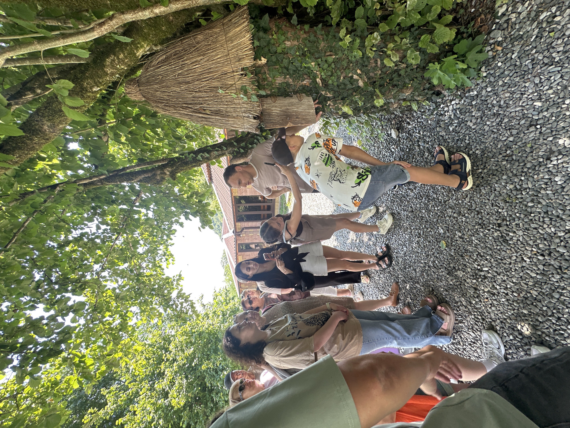 In Bzypta, Abkhazia, a group of people are gathered around a beehive constructed on a tree trunk, which is covered in ivy. Sunlight filters through the lush green canopy of trees, dappling the gravelly ground. A man, wearing a light brown t-shirt, stands and gestures towards the hive, while a young boy in a white t-shirt with a graphic print and denim shorts leans in to observe. A young girl in a brown dress stands next to the boy, also looking at the hive. Several other women and a man stand behind them, some holding phones. In the background, a building with a red tiled roof is visible through the trees. The overall scene depicts an educational or observational outing related to beekeeping in a natural setting.