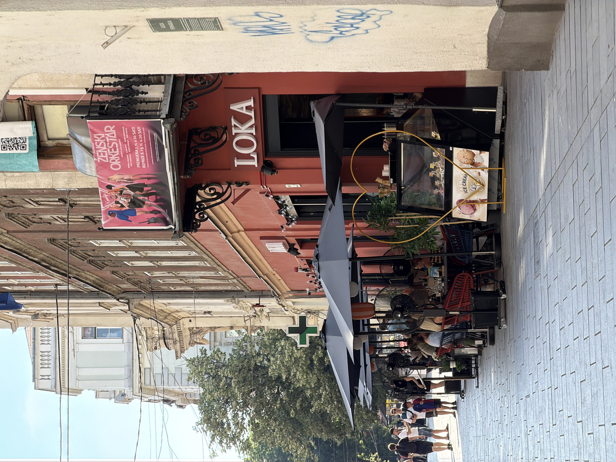 In Sarajevo, Bosnia and Herzegovina, a street-level outdoor seating area for a cafe or restaurant is captured on a sunny day. The foreground is a paved sidewalk made of light grey rectangular stones. To the right, a cream-colored building wall features prominent blue graffiti.
The main subject is a row of outdoor seating beneath dark grey umbrellas, occupied by several patrons. Fans are positioned around the seating area to provide cooling. In front of the establishment, a display case for ice cream is visible, adorned with a heart-shaped yellow frame. A sign in front of the display case advertises "PREMIUM ICE CREAM."
The building facade is painted a vibrant red and features an entrance with a sign that reads "LOKA" in white letters. Above the entrance, a balcony railing with decorative metalwork is present. A large banner advertising a "ŽENSKI ORKESTAR" (Women's Orchestra) is displayed on the building, along with a QR code.
Further down the street, trees and other buildings are visible. A green cross, typically signifying a pharmacy, is mounted on one of the buildings. Several people are walking along the sidewalk on the left side of the image, some appearing to be tourists. The overall atmosphere suggests a lively urban environment on a warm day.