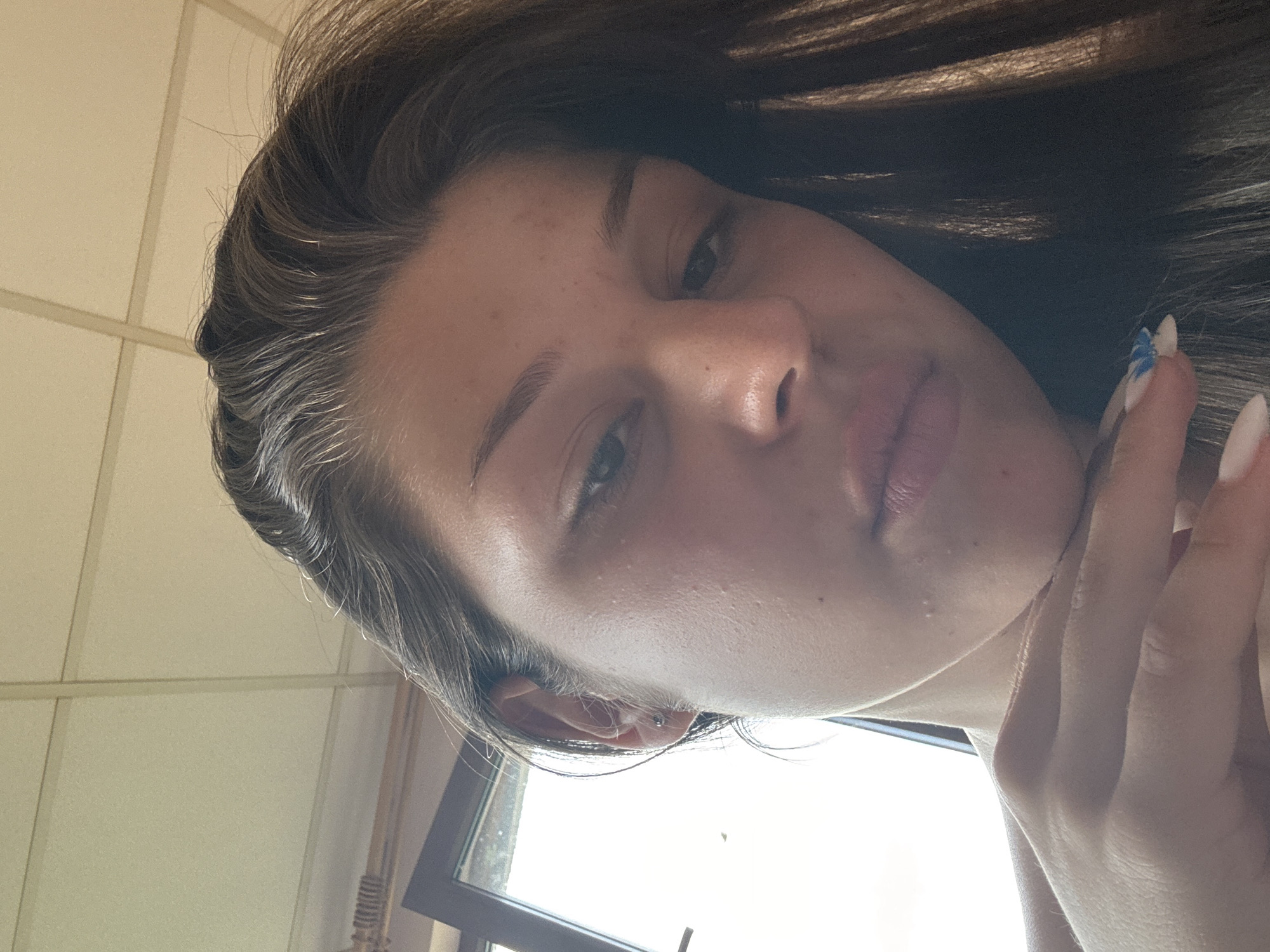 This is a close-up, rotated selfie of a young person. The subject has dark brown hair, fair skin with some light freckling, and is looking towards the camera. Their left eye is visible, with dark eyebrows and eyelashes. Their lips are slightly parted, and their right hand, with long fingernails painted with blue and white designs, is touching their face near their mouth. A small earring is visible in their left ear.
The background is out of focus but appears to be an indoor setting with cream-colored walls and what looks like a window or a door frame. The lighting suggests it could be daytime, with natural light illuminating the scene. The overall mood is casual and intimate, typical of a personal selfie. There is no visible text in the image.