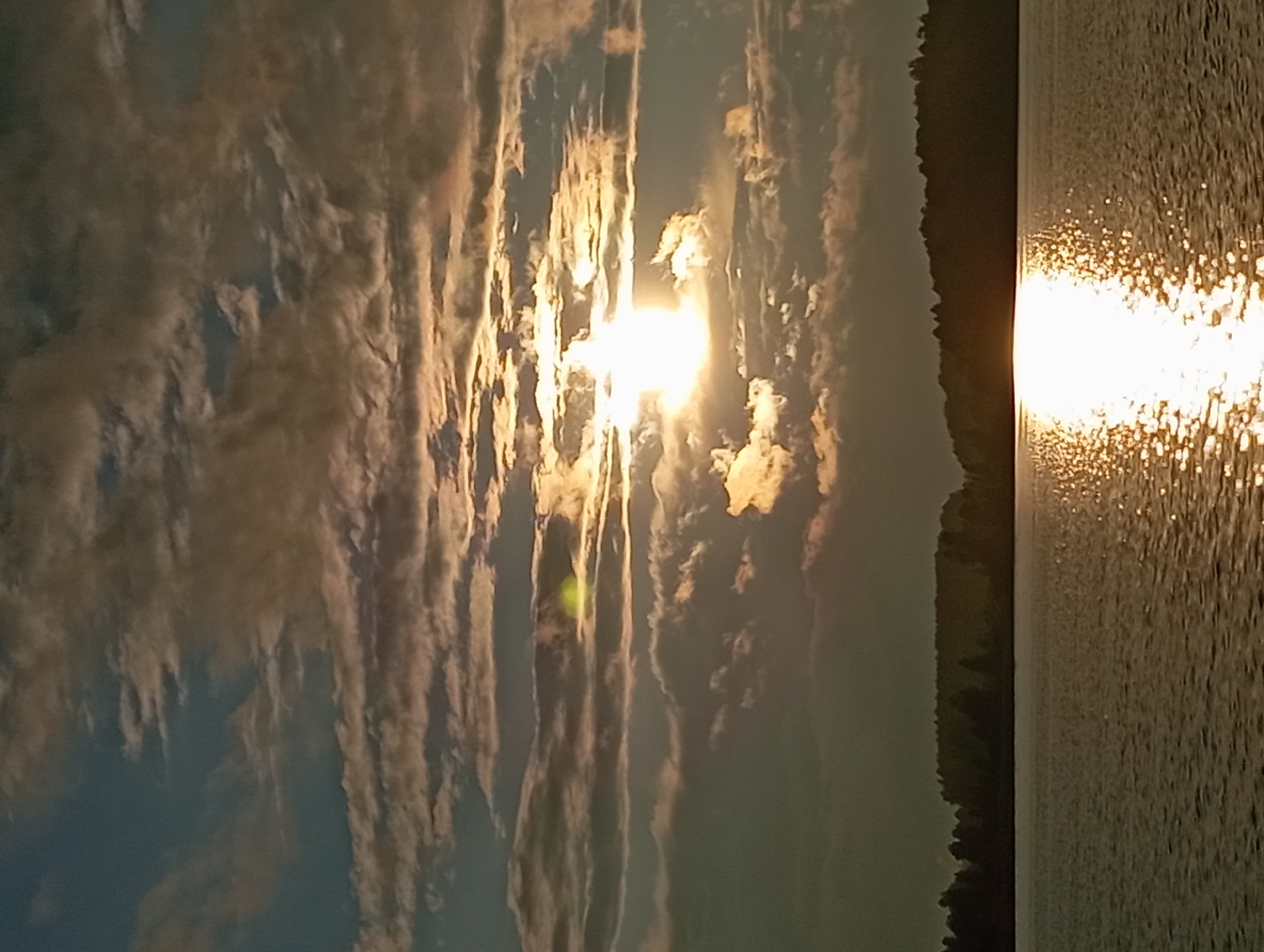 This is a horizontal, eye-level photograph of a lake and sky during sunset. The sun is low in the sky, partially obscured by clouds, casting a bright reflection on the water. The water is a muted brown-grey, with ripples catching the sunlight and creating a glittering effect. A dark, tree-lined silhouette of land stretches across the horizon, dividing the water and sky. The sky is a mix of blue, grey, and orange clouds, with the sun appearing as a bright white light in the center of the frame. The overall mood is serene and peaceful. The image captures a natural landscape, likely in Mołowiste, Poland, given the geographical context. There are no people or specific man-made objects visible in the scene, focusing solely on the natural elements of a sunset over water.
