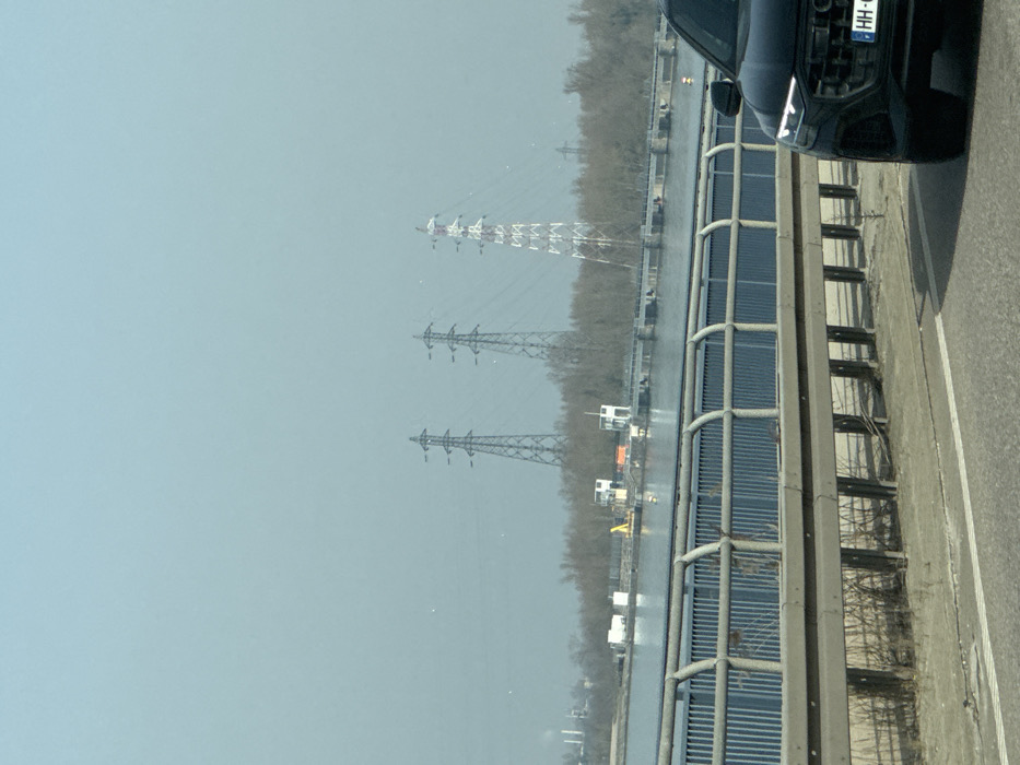 The image depicts a panoramic view from a moving vehicle on a bridge overlooking the Rhine River near Altenheim, Germany, under hazy conditions. In the immediate foreground, the front right section of a dark-colored SUV, bearing a German "HH-" license plate (Hamburg registration), is partially visible on the right. A gray metal guardrail and a blue metal barrier structure line the edge of the bridge. The midground features a wide body of water, likely the Rhine, with a dam or lock system visible across it, including multiple control buildings and industrial structures on the far bank. The distant background reveals a line of bare trees, above which three large lattice-type electricity transmission pylons are prominent. Two pylons are metallic grey, and the third (rightmost) is painted with alternating red and white bands. Further left in the background, industrial facilities with chimneys are faintly discernible. No individuals are present in the scene.