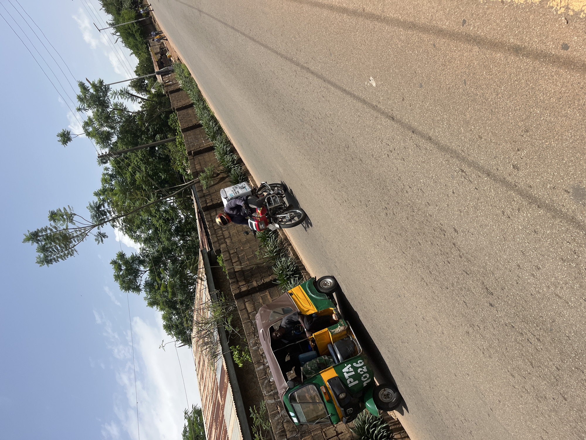 A person on a motorcycle drives down a paved road in Gura Top, Nigeria. The motorcycle is red and silver, and the rider is wearing a helmet and dark clothing. To the right of the motorcycle is a green and yellow auto-rickshaw, also known as a "Keke Napep," with three people inside. The auto-rickshaw has "PTA 1026" written on its side. Trees and a brick wall line the left side of the road, with some corrugated metal roofing visible behind the wall. The sky is blue with scattered clouds, suggesting it is daytime and the weather is pleasant. The overall scene depicts everyday transportation and urban life in Nigeria.