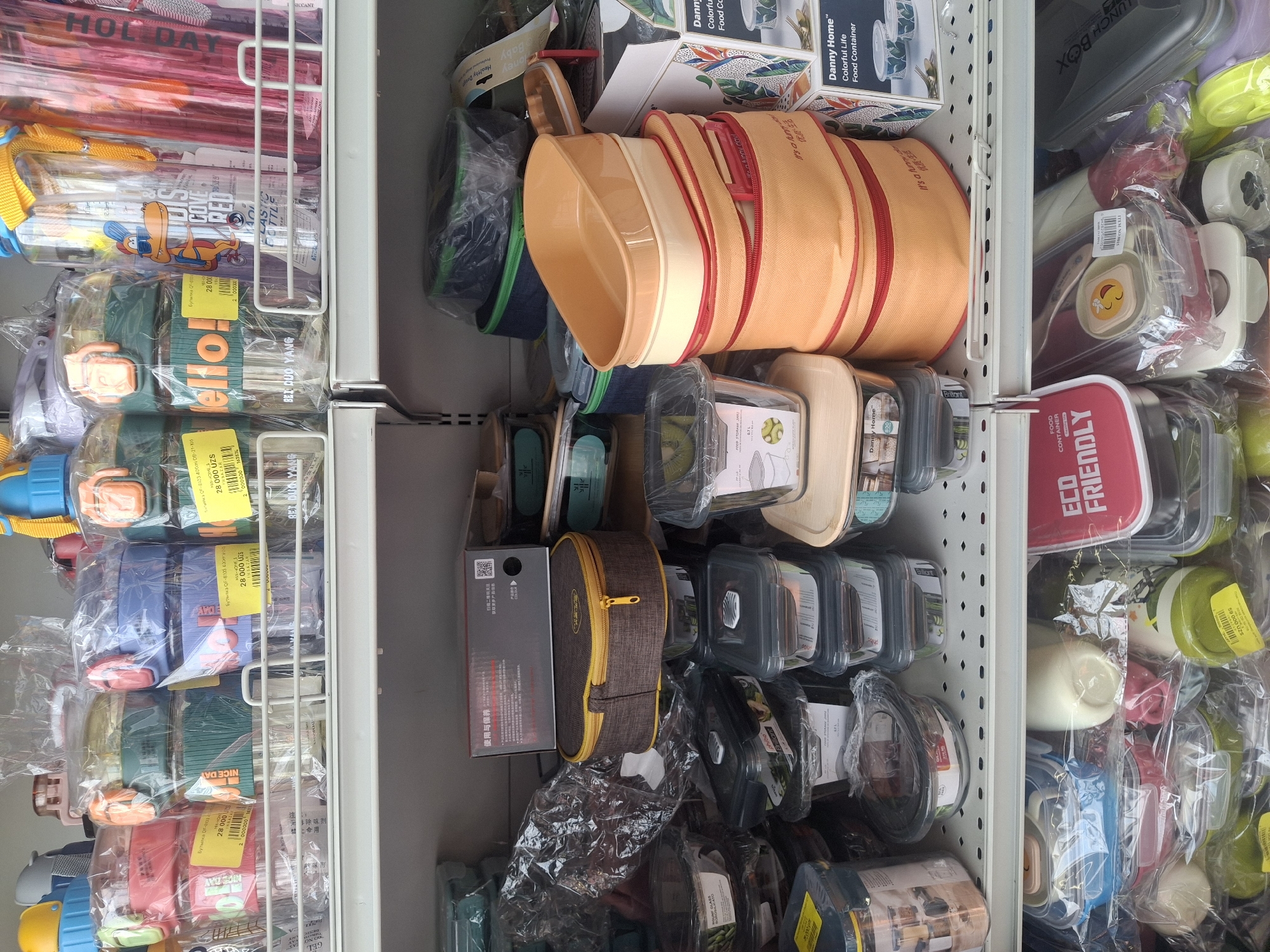 The image shows a retail display of food containers and bottles on shelves in a store. The products are mostly plastic and glass, with various colors and designs. Some items are packaged individually, while others are stacked. There are no people visible in the image, and no specific actions are taking place. The environment appears to be an indoor retail space, with shelves made of white metal. The lighting is artificial and consistent with typical store lighting. The image is taken from a medium distance, capturing a section of the store's inventory. The origin of the image is not explicitly stated, but the presence of products with Uzbekistani currency denominations (UZS) on some price tags suggests a location within Uzbekistan, potentially Tashkent.