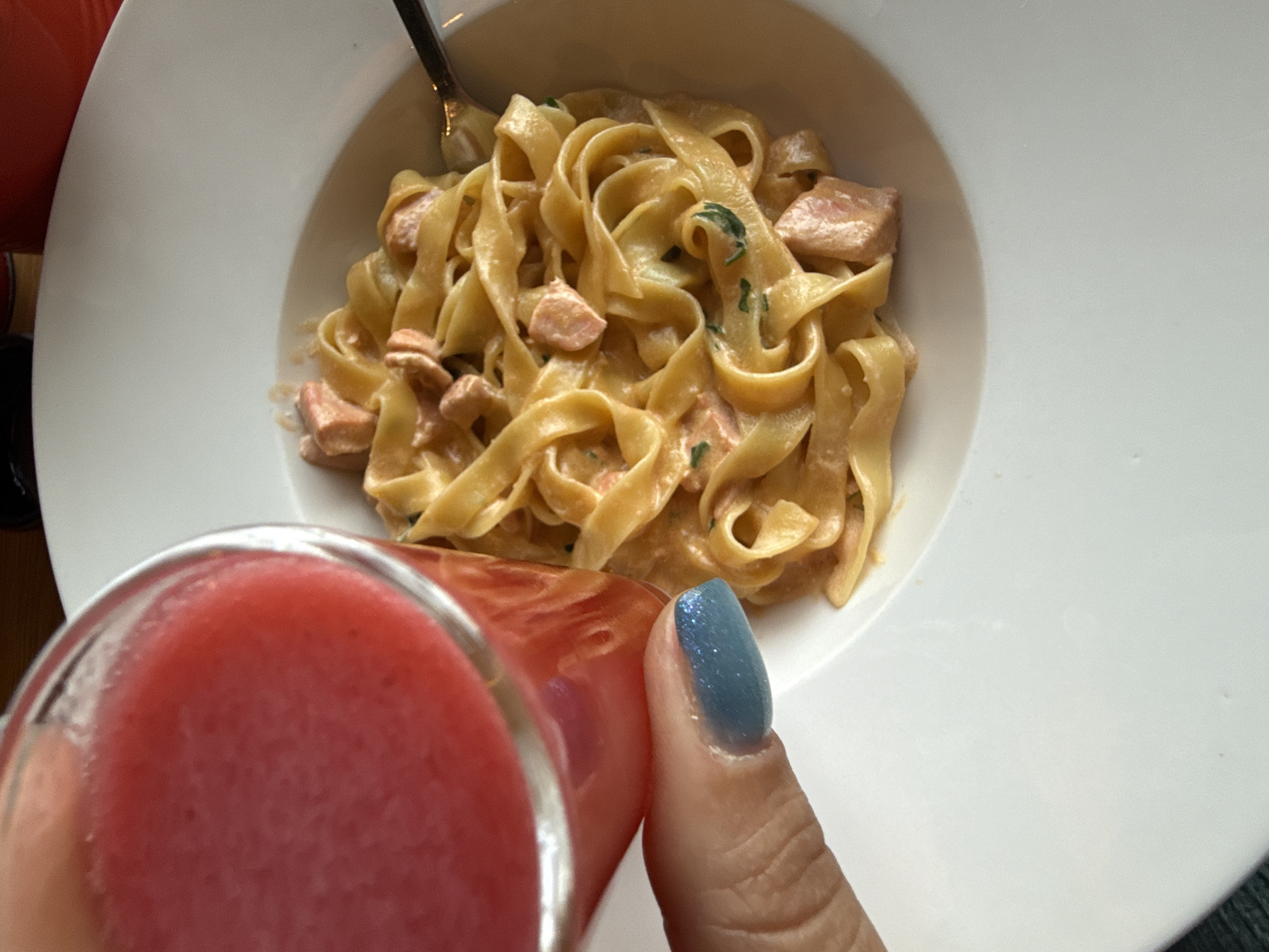 The image presents a close-up, top-down view of a meal and a drink, held by a person's hand, set in what appears to be a casual dining environment in Dmitrov, Russia.
The primary subject is a white, deep-rimmed plate filled with pasta. The pasta consists of broad, flat noodles, resembling tagliatelle or fettuccine, generously coated in a creamy, light orange-pink sauce. Numerous small, cooked pieces of protein, likely salmon or ham, are mixed throughout the pasta, along with visible flecks of green herbs, possibly parsley or basil. A silver fork is partially submerged in the pasta on the left side of the plate.
In the foreground, a clear glass contains a vibrant, opaque red beverage, which looks like a berry smoothie or juice. A person's hand is holding this glass, with the thumb and index finger clearly visible. The fingernail on the index finger is painted a striking, shimmery blue-teal color.
The scene captures the moment a person is about to enjoy their meal and drink. No visible text is present in the image. The time of day and specific emotions are not discernible.