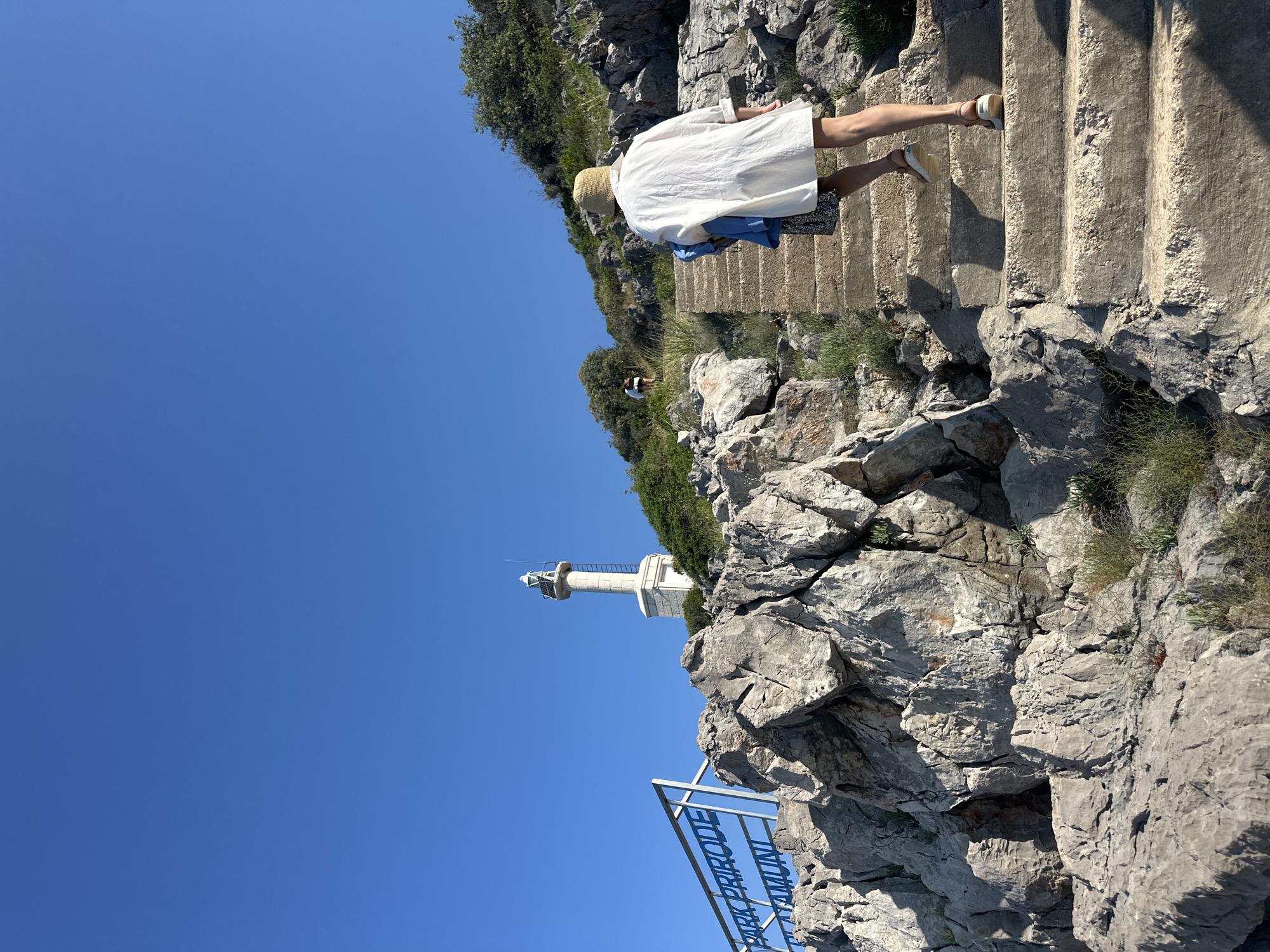 A person wearing a white shirt, straw hat, and sandals is walking up a concrete staircase on a rocky hillside under a clear blue sky. The person is carrying a blue garment. In the background, another person is visible further up the hill. A lighthouse stands on a higher elevation to the left, behind a rocky outcrop. A blue sign with white text, partially obscured by rocks, reads "PARK PRIRODE" and "ATAMUNI". The environment is characterized by rugged rocks, sparse vegetation, and the distant lighthouse. The scene is set in Prijevor, Montenegro.