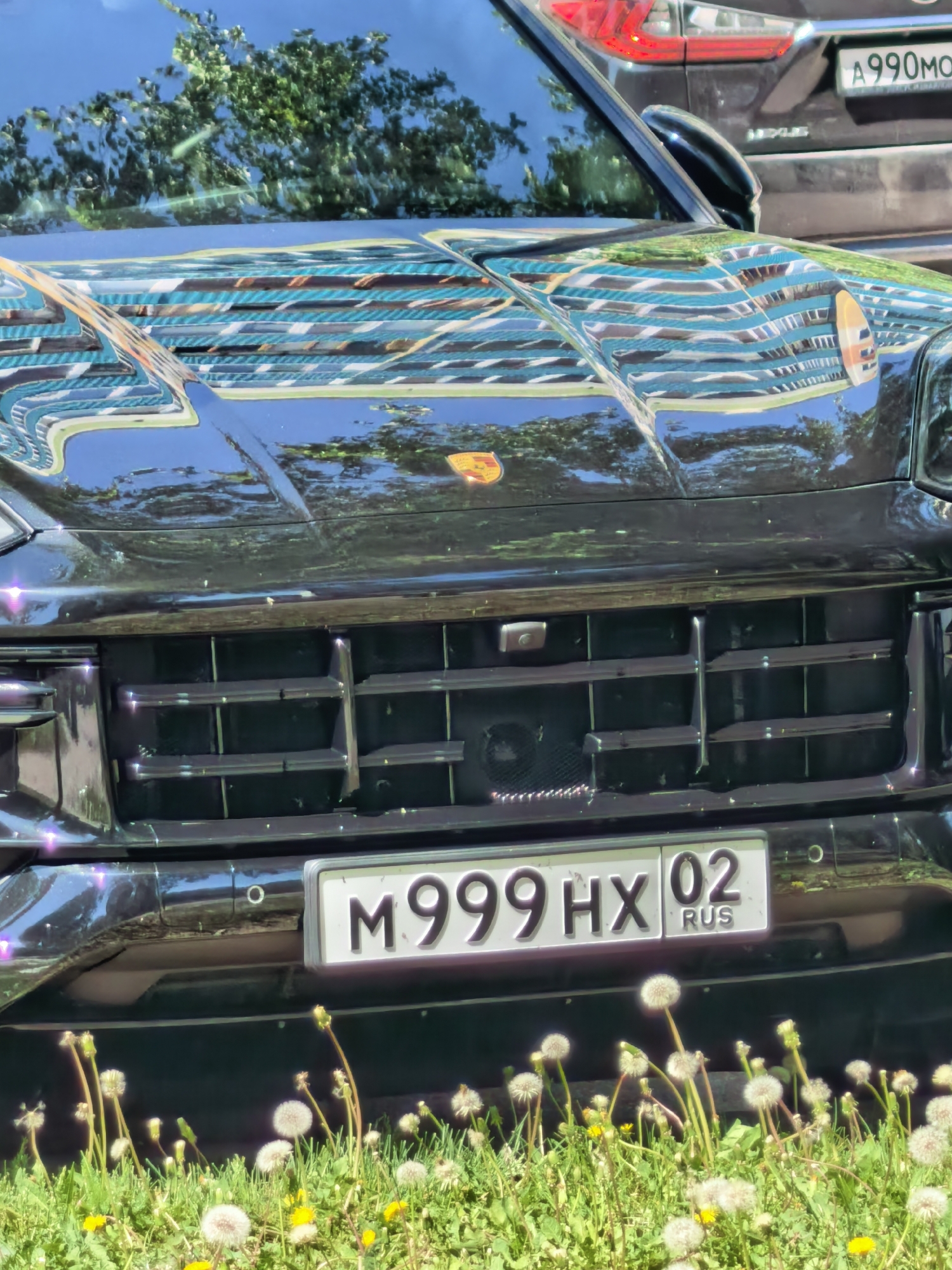 A black Porsche Macan is parked on a sunny day in Sterlitamak, Russia. The car has a Russian license plate that reads "M999HX 02 RUS". The front of the car reflects the surrounding buildings and trees. Dandelions grow in the grass in front of the car. A Lexus SUV is parked in the background with the partial license plate "A990MO".