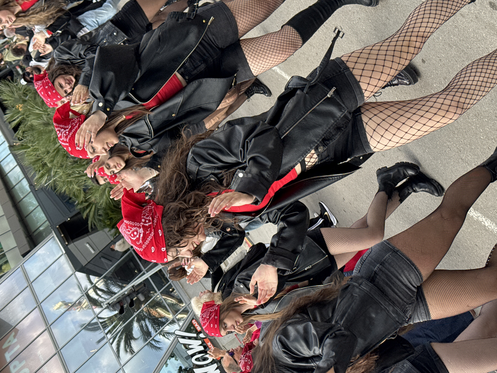 A group of people wearing black leather jackets and red bandanas are standing outside of a building in Budva, Montenegro. Most of the people appear to be wearing black shorts and fishnet stockings. The building is made of glass and has the words "JACK" and "BAR" written on it. There are palm trees in the background.