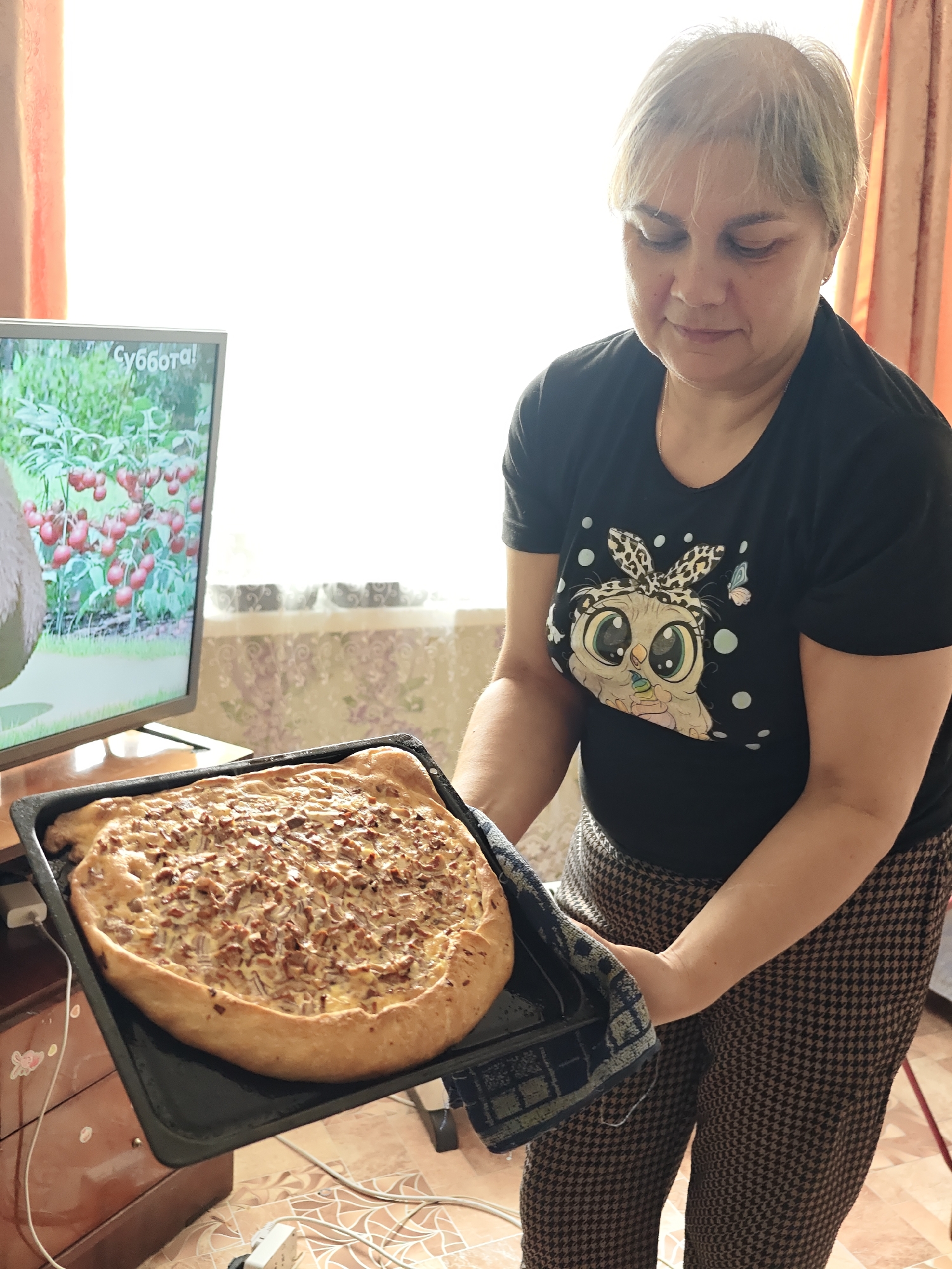 A woman in Уральское, Russia, holds a baking sheet with a freshly baked pie in her hands. She is wearing a black t-shirt with an owl print and patterned pants. A television in the background displays an image with the word "Суббота!" written on it. The room is brightly lit, suggesting it is daytime.