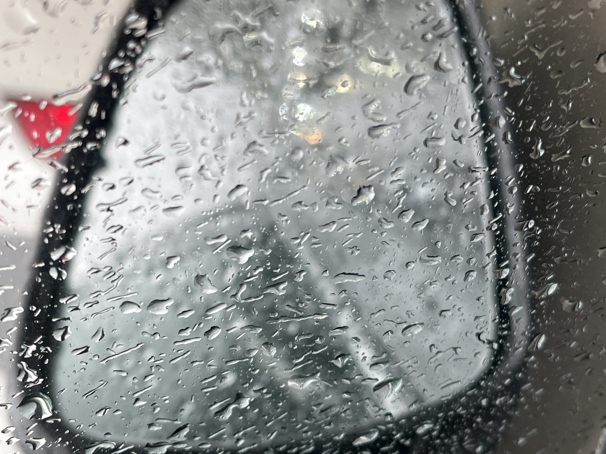The image is a close-up, high-angle shot of a car's side-view mirror covered in raindrops. The mirror reflects a blurry, desaturated scene of what appears to be a winter day, possibly in Nizhny Novgorod, Russia. The weather is clearly inclement, with numerous water droplets distorting the view.
The foreground is dominated by the texture of the raindrops on the glass, creating abstract patterns and highlighting the surface. Some drops are small and rounded, while others have elongated and streaked, suggesting movement or wind. The mirror itself is dark and framed by a black edge.
In the reflection, hints of the external environment are visible but heavily obscured by the water. There are blurred shapes that could be trees or buildings, rendered in shades of grey and white. A bright red triangular object, possibly a warning sign or part of another vehicle, is visible on the left side of the frame, its color muted by the rain. Some faint, blurry lights, possibly headlights or streetlights, are also discernible in the distance, indicating it might be daytime or early evening.
The overall mood is one of enclosure and perhaps a slight sense of isolation due to the weather. The scene suggests the experience of being inside a vehicle during a rainy or snowy day, with the mirror offering a fragmented view of the outside world. There is no visible text in the image.