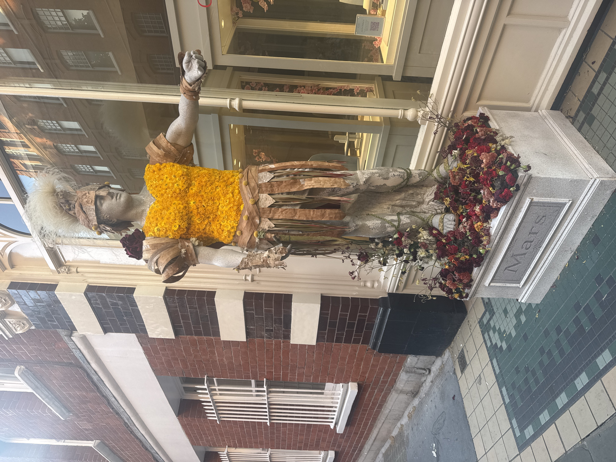 A statue titled "Mars" stands on a city street. The statue is made of stone or a stone-like material. It wears a helmet adorned with white feathers and a bright yellow flower chest plate. Dried leaves are wrapped around its body. It is surrounded by dried red flowers. The statue stands in front of a storefront with a glass window reflecting a building across the street. The building next to the store is brick with white and black paint above it. The sidewalk is made of green and white mosaic tiles. The city is unknown.