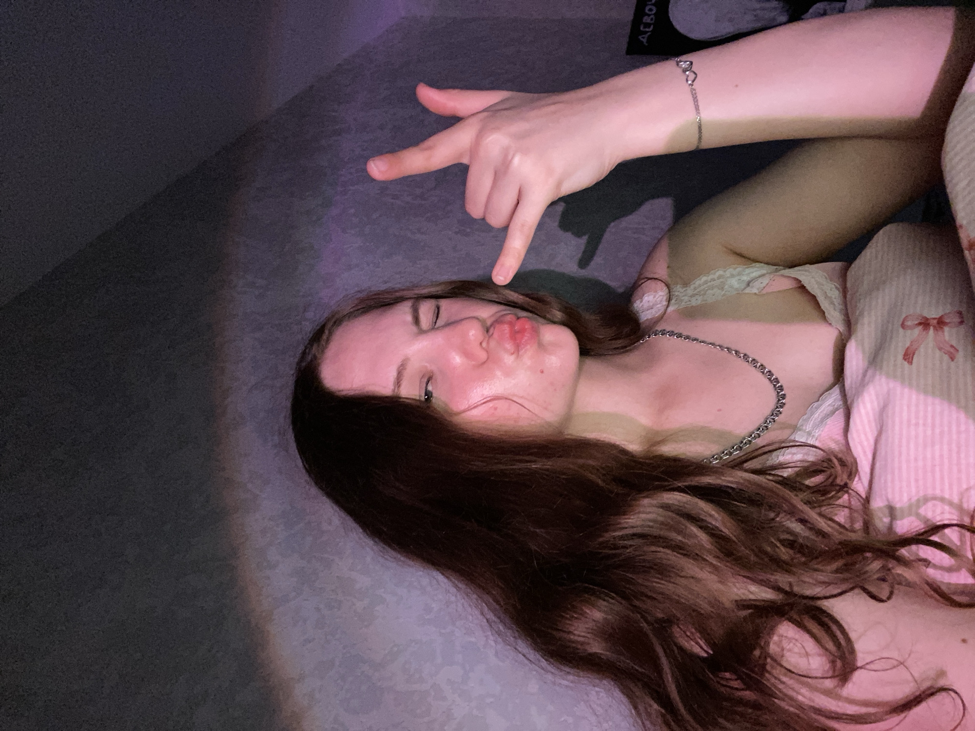 A young woman with long, dark brown hair is captured in a close-up, self-taken photograph, likely indoors. She is winking and puckering her lips, making a "rock on" hand gesture with her right hand. She wears a silver chain necklace and a bracelet with heart-shaped charms on her left wrist. Her attire appears to be a pink and white striped garment, possibly pajamas or a camisole with lace trim. The background consists of a textured wall with a gradient of purple and pink lighting, suggesting ambient colored light. The lighting creates a shadow of her hand on the wall. No specific details place this image within the city of Izhevsk, Russia.