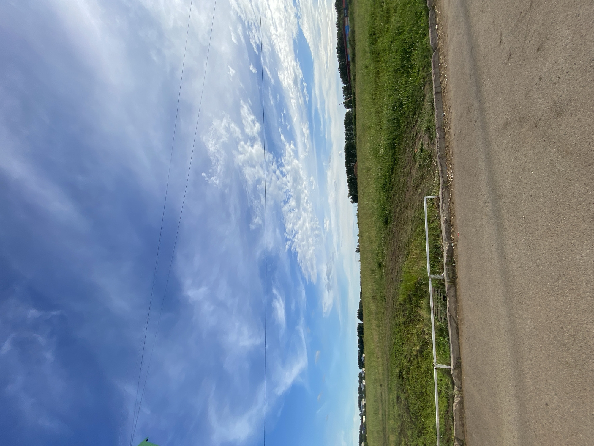 The image captures a rural or suburban landscape under a bright blue sky with scattered white clouds. In the foreground, a paved road runs horizontally across the bottom of the frame, with a concrete curb and a low white metal fence separating it from a grassy field. The field is covered in lush green grass and appears somewhat uneven. In the middle ground, a line of trees forms a dark horizontal band, with a few scattered buildings visible in the distance to the left, suggesting a settlement. The sky is expansive, with wispy white clouds streaking across the blue. There are no people or animals visible in the image. The scene appears to be an undeveloped or semi-developed area on the outskirts of a town or city. It is possible this is a landscape photograph taken in Dyurtyuli, Russia, given the context provided.