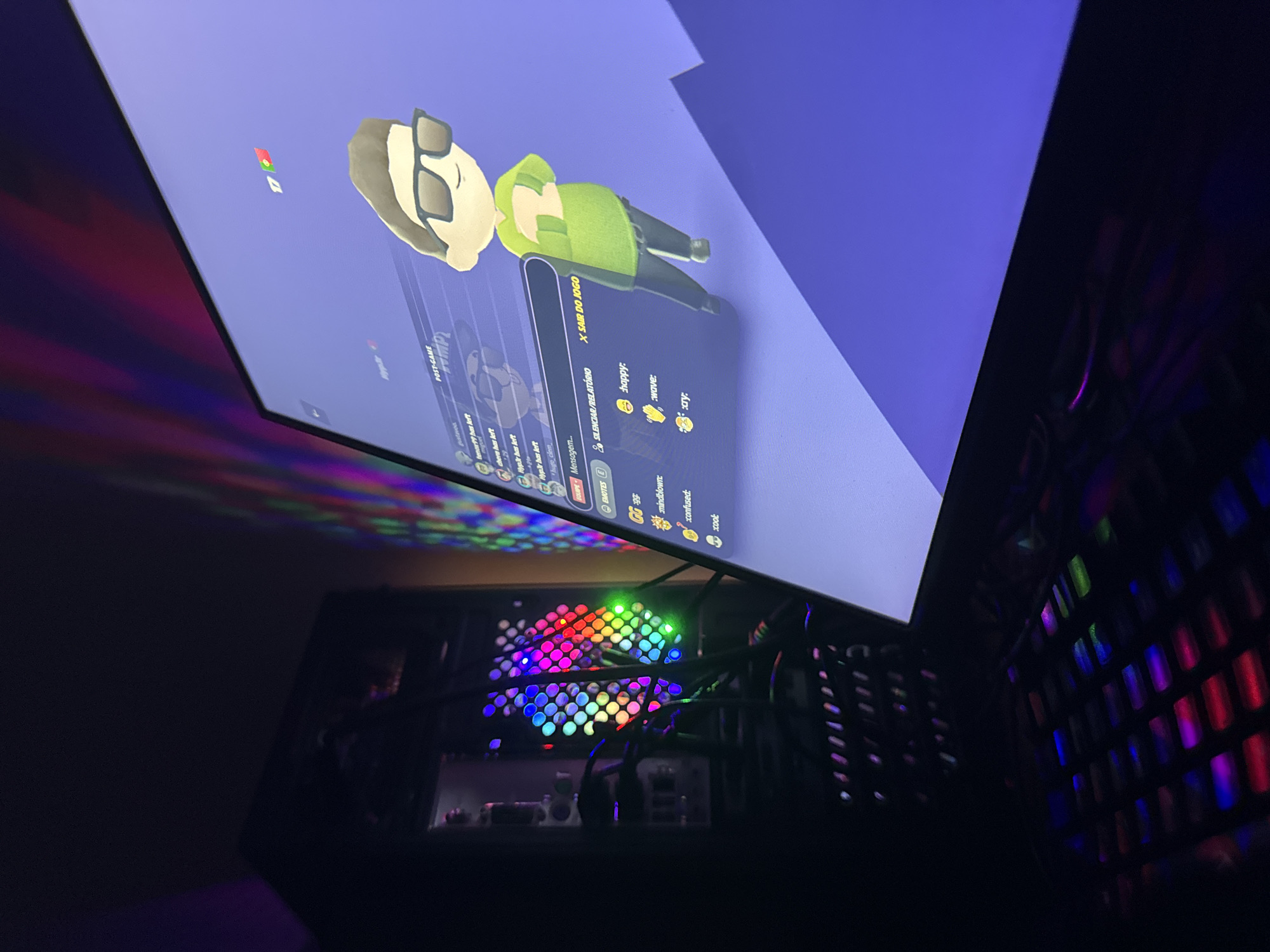 The image shows a computer setup in a dimly lit room. A monitor displays a screen capture from a video game or social platform featuring a cartoon avatar wearing sunglasses and a green outfit. The screen also contains text, indicating post-game options, chat messages, and emote selections.
Beneath the monitor is a partially visible computer case with colorful RGB lighting emanating from internal fans. The computer case appears to be black and has visible ventilation grills.
The surrounding environment is dark, suggesting an indoor space with limited lighting. There is no evidence in the image to place it in Mirandela, Portugal.