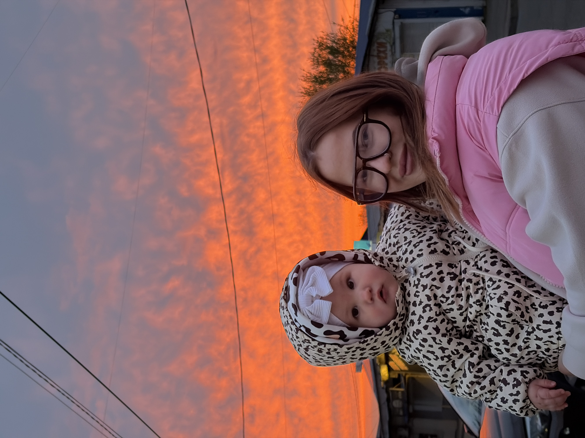 The image, oriented vertically, captures a young woman holding a baby outdoors against a vivid sunset sky, likely in an urban setting like Krasnodar, Russia.
The upper portion of the frame is dominated by a spectacular sky displaying a gradient of intense orange and reddish hues, transitioning to softer purples and blues. Scattered clouds catch the last light of the day, creating a dramatic backdrop. Several dark, thin power lines stretch across the sky.
In the foreground, a young woman with light brown hair and black-rimmed glasses looks directly at the camera with a calm, subtle smile. She wears a bright pink puffer vest over a light-colored hooded sweatshirt and has a nose piercing. She holds a baby, who also faces the camera with wide, curious eyes. The baby is warmly dressed in a hooded leopard-print puffer jacket, with a white, ribbed bow visible on its head beneath the hood.
The background shows faint, blurred elements of an urban environment, including distant buildings and sparse foliage from a tree. On one of the distant buildings, there appears to be some indistinct Cyrillic text. The overall scene conveys a warm and serene atmosphere, illuminated by the striking natural light of the sunset.