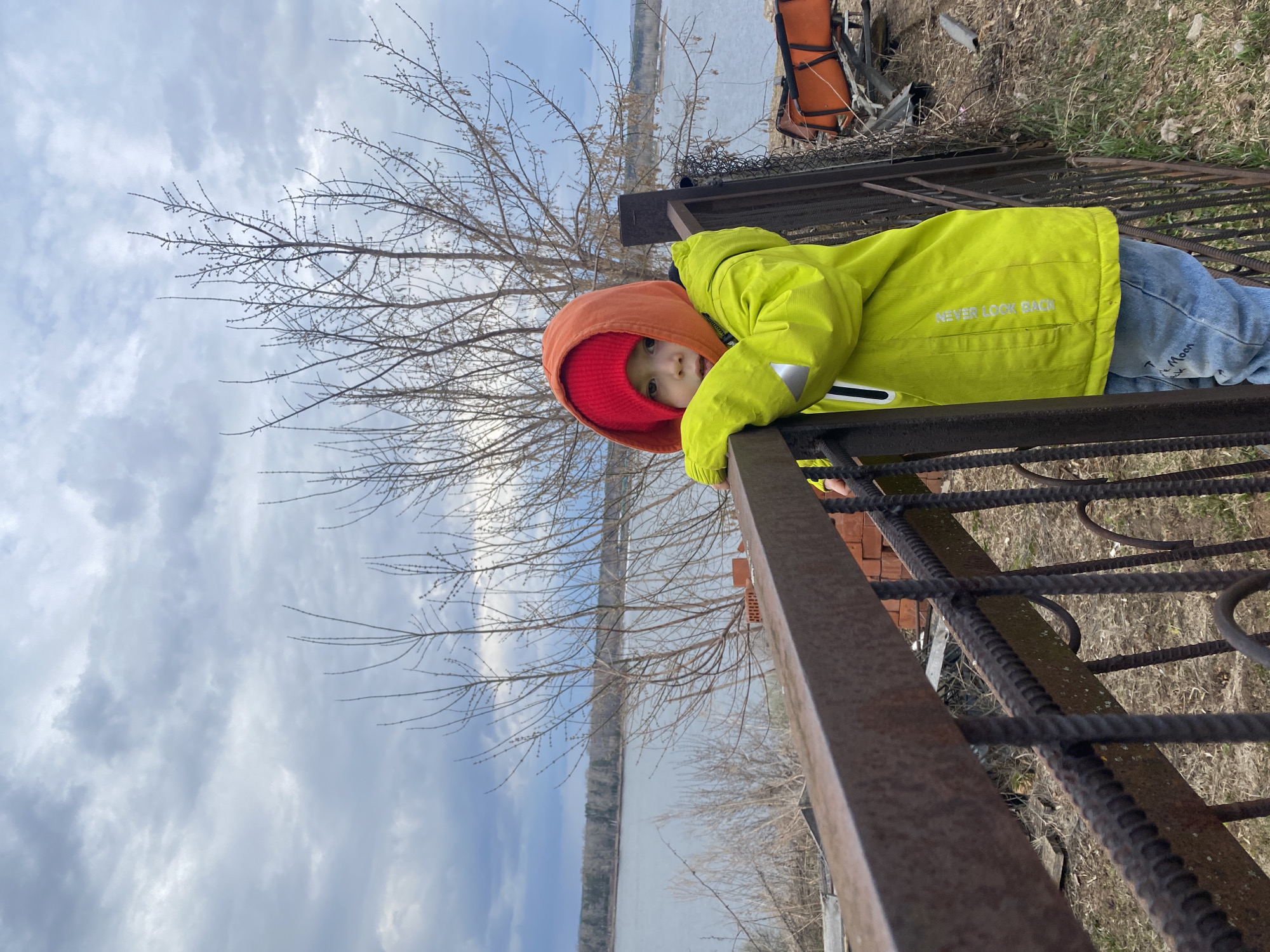 A young child, wearing a lime green jacket with the words "NEVER LOOK BACK" on the back, an orange hood, and a red hat, is leaning over a rusted metal fence. The child is looking towards the camera. Behind the fence is a body of water, with trees lining the opposite shore, and a cloudy sky above. An orange life raft is partially visible on top of a brick structure behind the fence. The location is Ольховка, Russia.