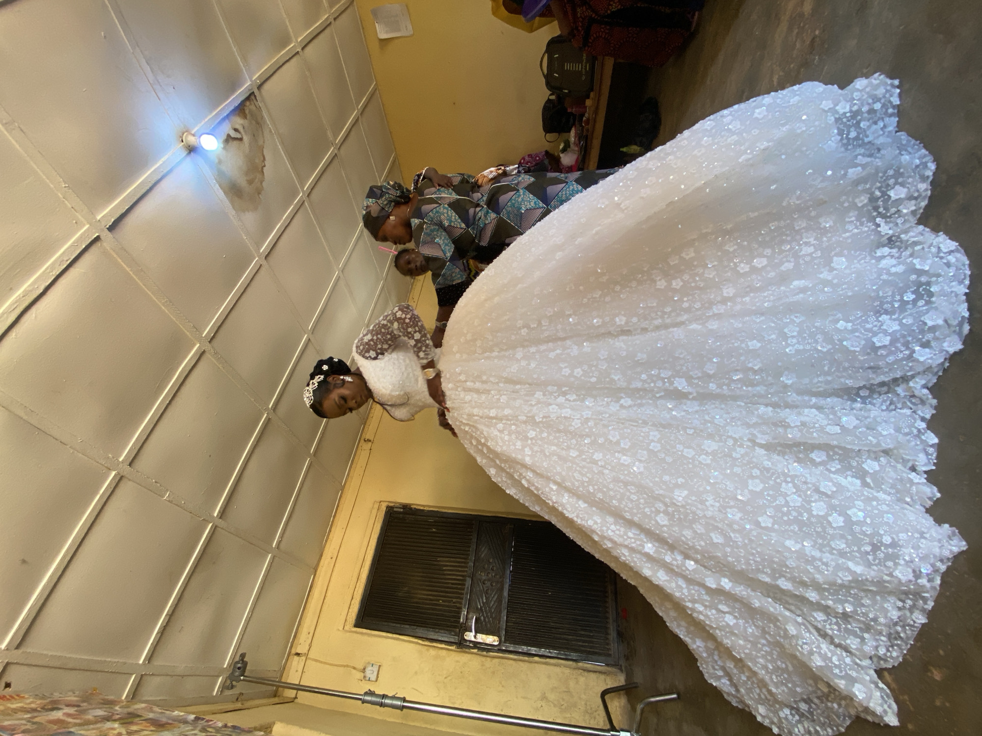 The image depicts a bride in Vom, Nigeria, wearing a white wedding gown with floral embellishments, attended by two people. The location appears to be indoors, with a yellowish wall and a window with closed shutters. The room has a grid-patterned ceiling with a visible light fixture. The scene shows the bride being attended to, likely preparing for a wedding ceremony. The environment is simple, with minimal decorations visible. A luggage bag and other items are on a shelf in the background.