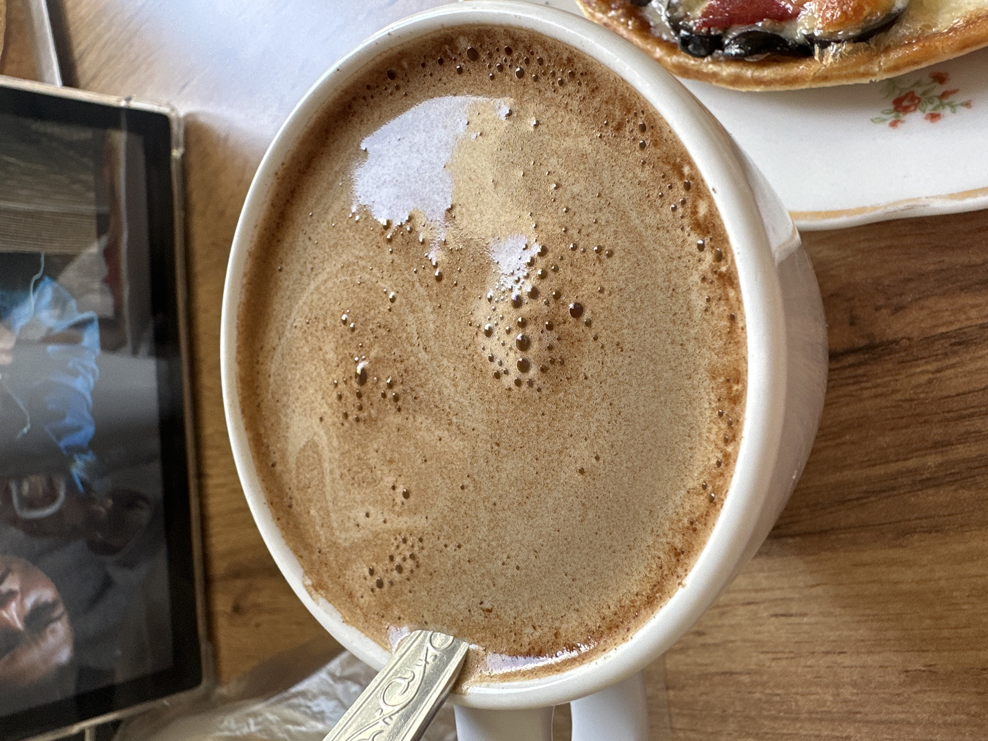 A close-up, overhead shot reveals a table setting with a warm beverage, a pastry, and a tablet, suggesting a casual break or meal. The central focus is a white ceramic cup filled with a light brown, frothy drink, likely coffee or hot chocolate, with numerous small bubbles on its surface. A silver spoon with an ornate, possibly floral, embossed design is partially submerged in the beverage.
To the left, a black-framed tablet rests on the wooden table. Its screen displays a reflection, within which a blurred image of a person's face and a blue garment are discernable, indicating someone is present or nearby. On the upper right, a portion of a white plate with a delicate floral pattern (small red and green flowers) and a gold rim is visible. Resting on this plate is a pastry, possibly a tart or danish, topped with dark elements (perhaps olives or berries) and reddish ingredients over a lighter base. All items are placed on a wooden surface, showing distinct grain patterns.
Bright reflections on the drink's surface and the tablet suggest ample natural light, indicating daytime. The scene evokes a sense of calm and relaxation, consistent with a coffee break or light meal. While the context places the image in Stavropol, Russia, no specific visual cues within the photo confirm this. No text is visible in the image.