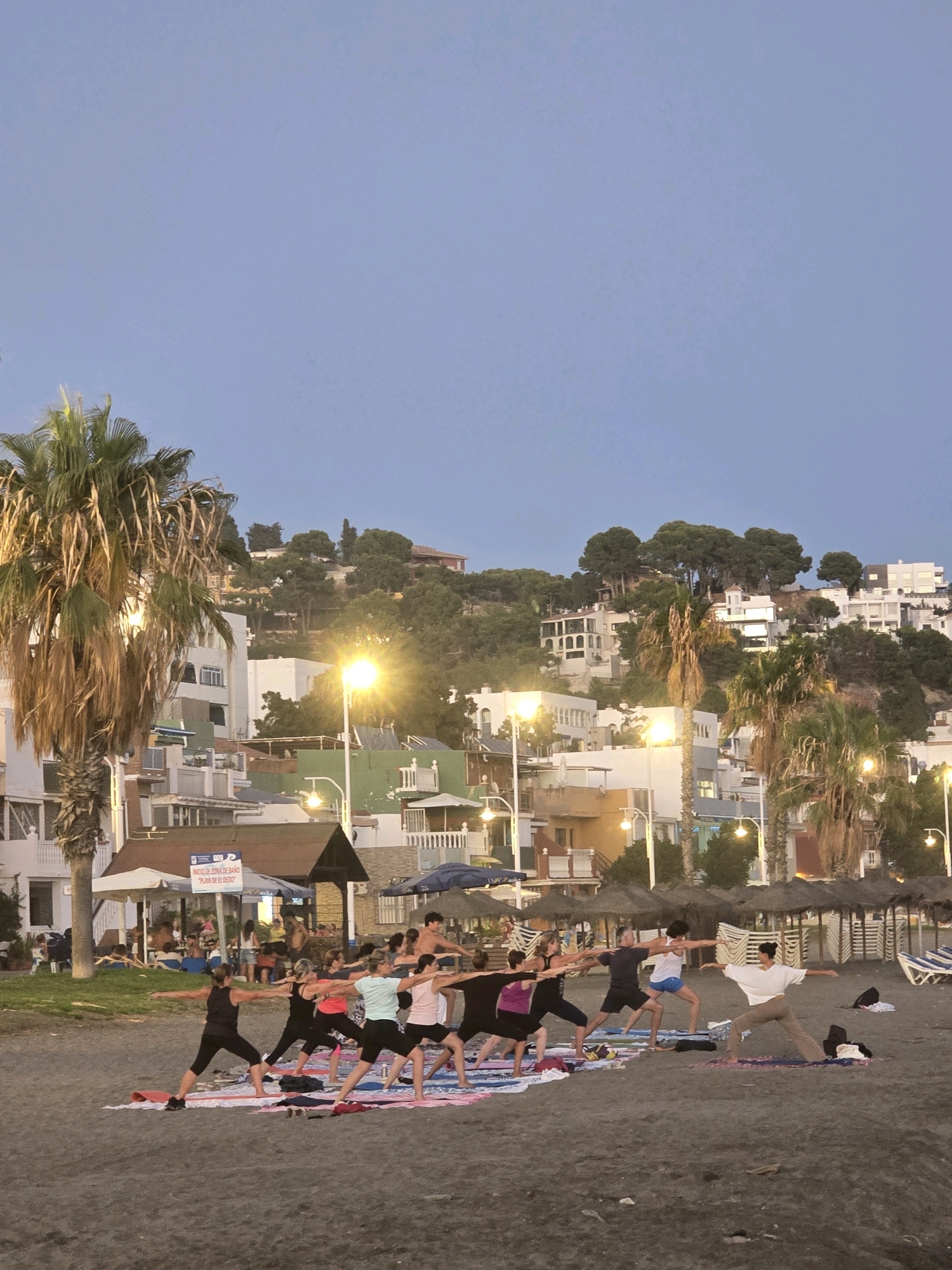 This is a medium shot, eye-level photograph of a group of people practicing yoga on a beach at dusk, in Málaga, Spain. The sky is a pale blue, transitioning into a darker blue at the top of the frame. In the foreground, a group of approximately 10 individuals are arranged in a line, facing towards the left of the frame, performing a yoga pose that involves outstretched arms and legs in a wide stance. They are positioned on individual yoga mats spread across the dark sand of the beach.
The individuals are diverse in age and gender. They are wearing athletic clothing in various colors, including black, pink, white, and blue. Some participants are looking forward, while others are looking towards their outstretched hands.
Behind the yoga group, there is a slightly elevated area with buildings that appear to be hotels or apartments, stacked on a hillside. These buildings are whitewashed with some colored accents in green and orange. Several palm trees are visible, with one large palm tree prominently placed on the left side of the frame. Streetlights are illuminated throughout the scene, casting a warm glow. In the background, further up the hill, more buildings and trees are visible. The beach itself is composed of dark sand, and in the distance, beach umbrellas and lounge chairs are set up, some of which appear to be folded or covered. A sign in Spanish with "INICIO DE ZONA DE BAÑO" and "PLAYA DE EL DEDO" is visible on the left side of the frame, indicating this is the beginning of the bathing zone for El Dedo Beach. The overall atmosphere is serene and active, with the group engaged in a communal exercise session as the day winds down.