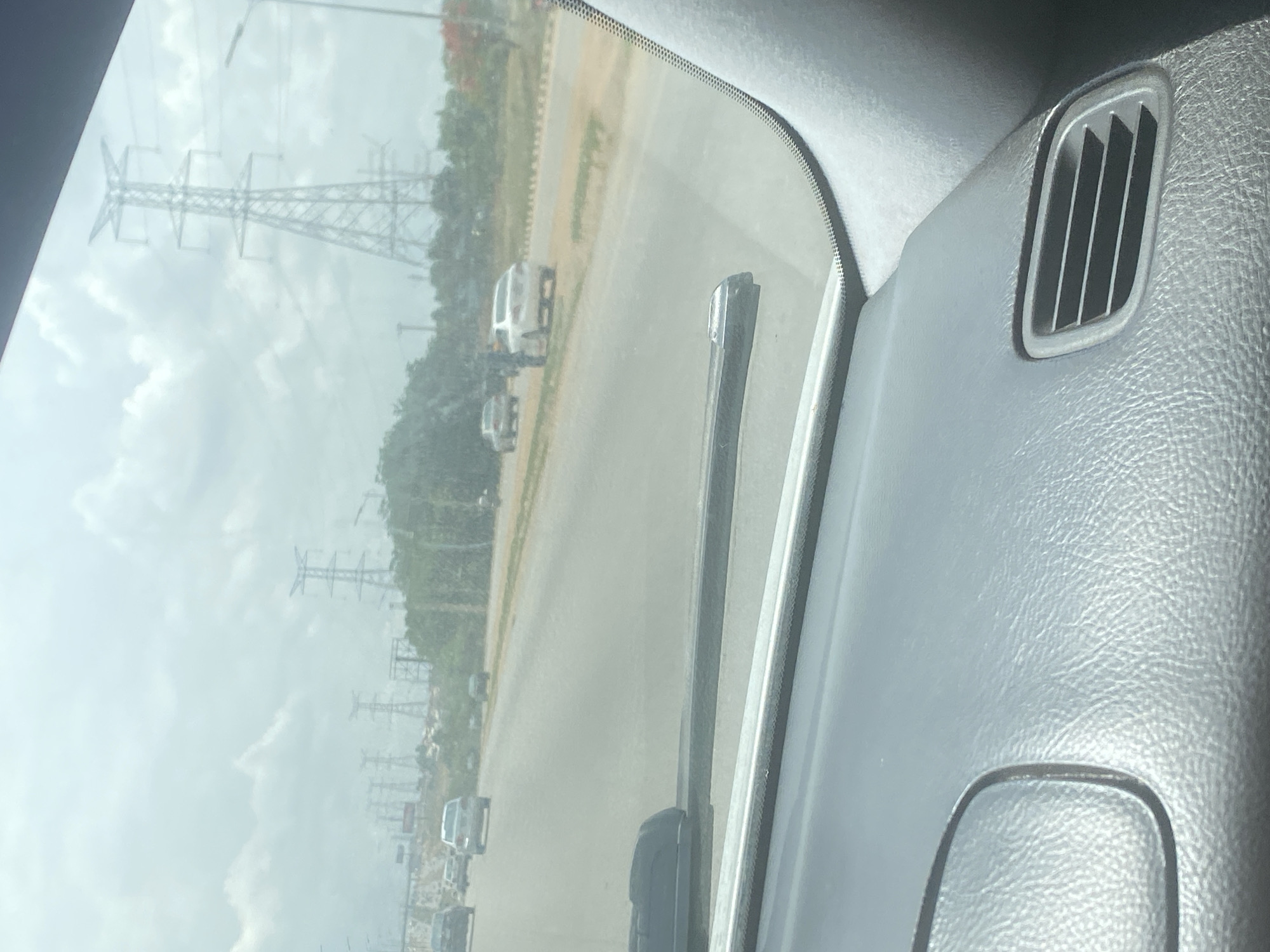 The image depicts a view from inside a vehicle, likely a car, looking out through the windshield. The scene outside the windshield shows a road with several vehicles in the distance. Along the side of the road are electrical transmission towers. The sky is overcast. Inside the vehicle, part of the dashboard and an air vent are visible. The location is inferred to be in or near Iddo Tudunwada, Nigeria, based on the prompt's instructions, although there are no distinctive features visible in the image to confirm this.
