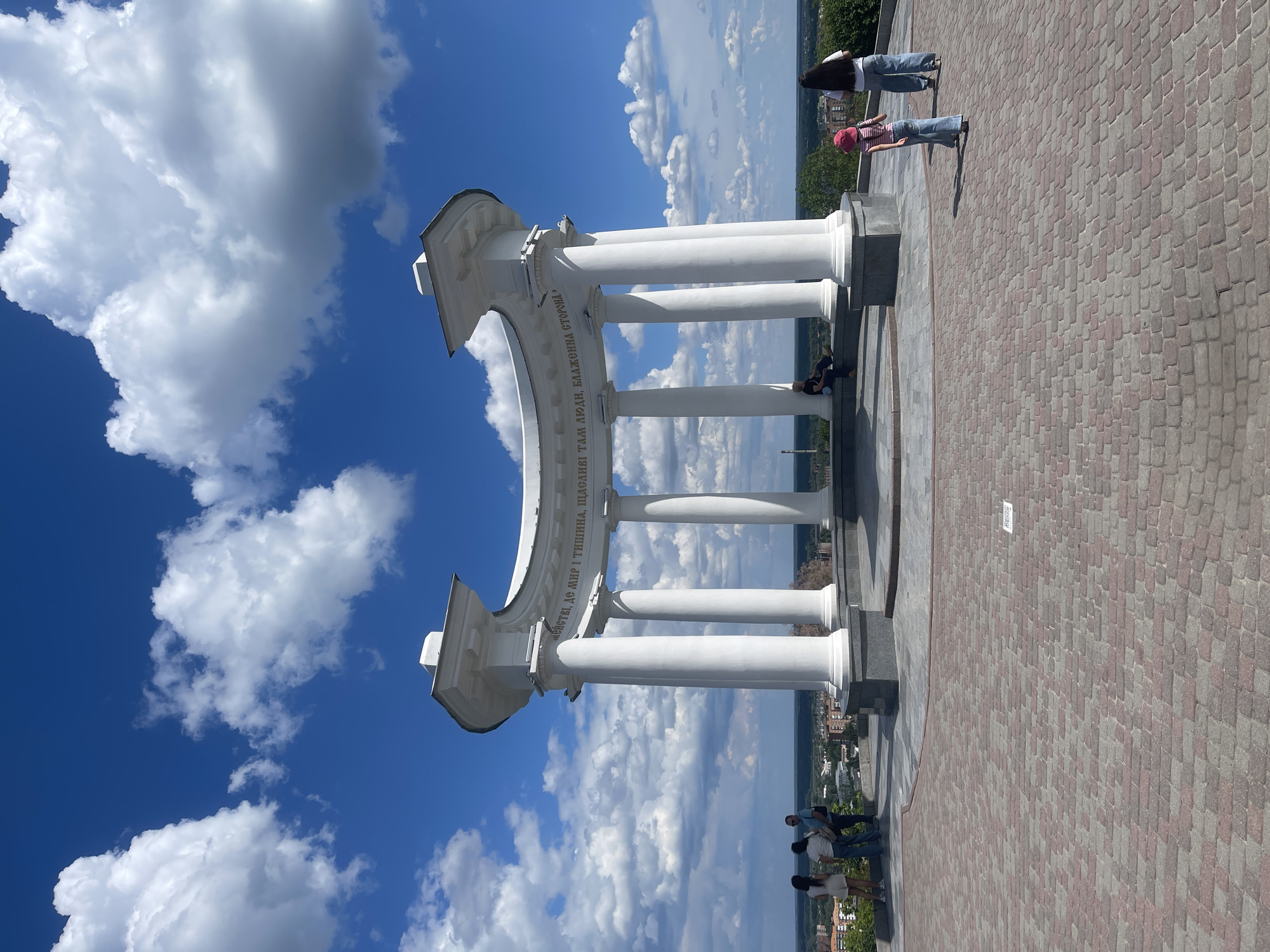 The image captures a vibrant, sunlit daytime scene at a scenic overlook, centered around a large, white, classical-style archway. The sky is a brilliant blue, adorned with numerous large, puffy white cumulus clouds, suggesting fair weather.
The main subject is a substantial semicircular arch, supported by several tall, white columns, likely made of stone or concrete. Golden lettering is inscribed along the inner curve of the arch. The visible text, transcribed as "мейства, де мИР І ТИШИНА, ЩАСЛИВІ ТАМ ЛЮДИ, БЛАЖЕННА СТОРОНА," is likely Ukrainian or Russian, translating approximately to "family, where there is peace and quiet, happy are the people there, blessed is the side/land."
The arch rests on a wide paved area, characterized by interlocking reddish and grey bricks. Several people are present, enjoying the outdoor setting. On the right side of the pavement, two women stand, one with long dark hair and jeans, and the other wearing a red hat and a striped shirt. Underneath the arch, on the left, two individuals are casually seated at the base of a column. Further to the left on the paved area, a group of three people, including a man and two women, are standing.
In the background, beyond the paved area and the arch, a vast expanse of distant landscape is visible, featuring what appears to be a city or town with buildings and abundant green foliage, indicative of a view from a significant elevation. The overall impression is one of a relaxed, pleasant day, with people enjoying the panoramic views and the architectural monument.