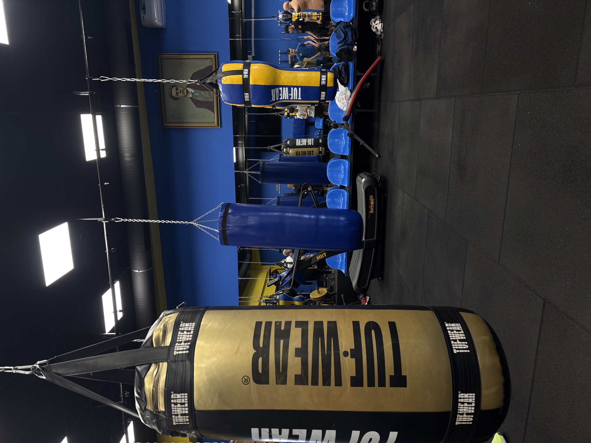 This indoor shot shows a gym setting in Sofia, Bulgaria, with several hanging boxing bags prominently displayed. The bags bear the brand name "TUF-WEAR". In the background, people can be seen engaging in various fitness activities, including boxing and using exercise machines. The room has a color scheme of blue and black. There are blue chairs lined up in rows, and a portrait hangs on the wall. Exercise equipment, including a "ReNegaDE" machine, is visible.