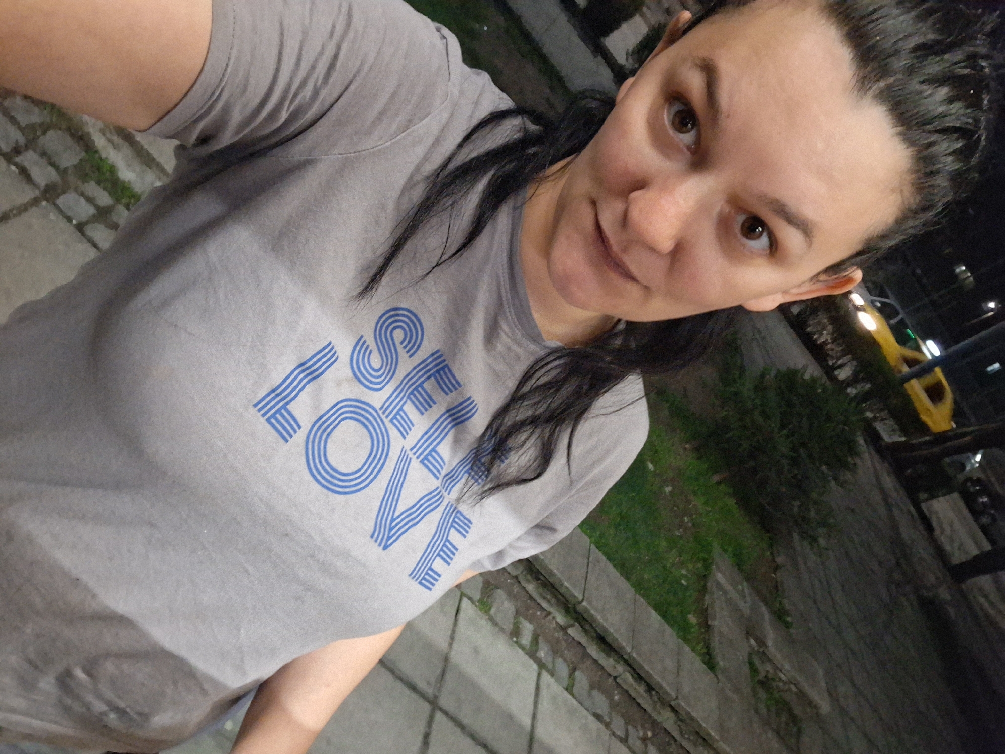A selfie of a woman is centered in the frame, taken outside in a possible city location. The woman has dark hair pulled back, brown eyes, and is wearing a gray T-shirt with the word "LOVE" in blue letters on it. The lighting is dim, suggesting it is evening or night. In the background, there is greenery and a glimpse of what appears to be a yellow vehicle, possibly a car or taxi. The ground is paved, and there are architectural elements like walls or curbs visible. Based on the prompt the location is Goce Delcev, Bulgaria, though this is not visually verifiable.
