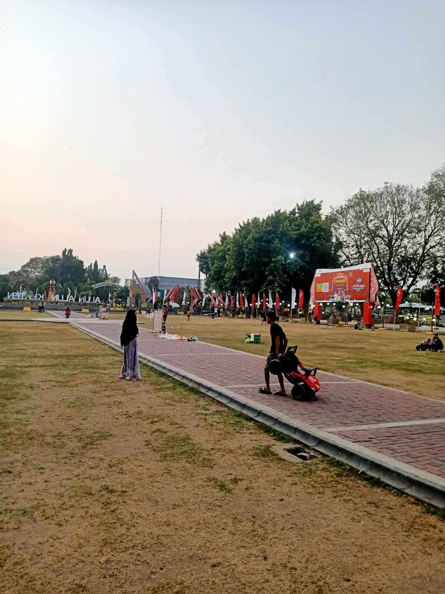This is a photograph taken at Alun-Alun Klaten, a public square in Klaten, Indonesia, during the late afternoon or early evening. The sky is clear and pale, suggesting the sun is low.
In the foreground, a paved pathway with red brick pavers runs diagonally across the image. On the left side of the pathway, a person wearing a black hijab and a light blue and grey patterned skirt stands facing away from the camera. To the right of the pathway, a young person in dark shorts and a black t-shirt is seen from the back, pushing a small red toy car. The ground around the pathway is covered in dry, yellowish-brown grass.
In the middle ground, the Alun-Alun is set up with various stalls and decorations. Several flags, some red and white, are visible. A large red banner with text and images is prominently displayed, possibly advertising an event. Trees form a backdrop behind the activity. In the distance, there are more trees and buildings.
The scene depicts a public gathering or event, with people milling about in the park area. The overall atmosphere appears calm and relaxed, characteristic of a public space in the evening.
