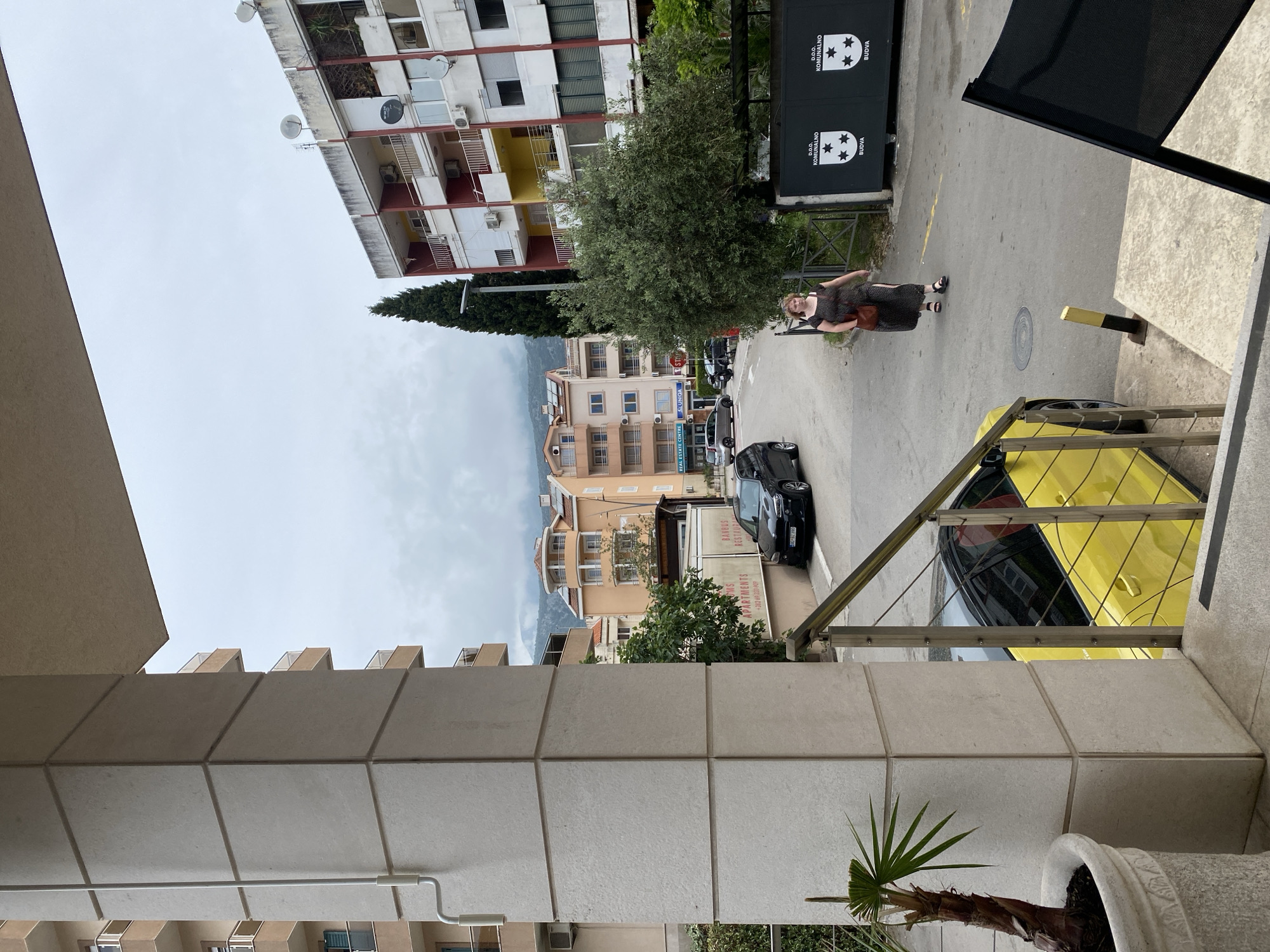 A daytime shot from an elevated vantage point shows a street scene in Budva, Montenegro. A woman in a dark dress and sandals walks along the street, carrying a brown bag. Behind her is a black sign with white lettering and the city name "BUDVA" along with stars. A black car is parked behind her.
Across the street are several buildings. The closest is a tan building with "BANBUS RESTAUR" visible on it. Further down are buildings in a tan and white palette, including one with red and yellow accents on the upper levels. The sky is overcast.
In the foreground, a balcony or terrace is visible. It's made of square, light-colored stone tiles. A small palm tree in a white pot stands to the right. A yellow car is partially visible behind the railing.