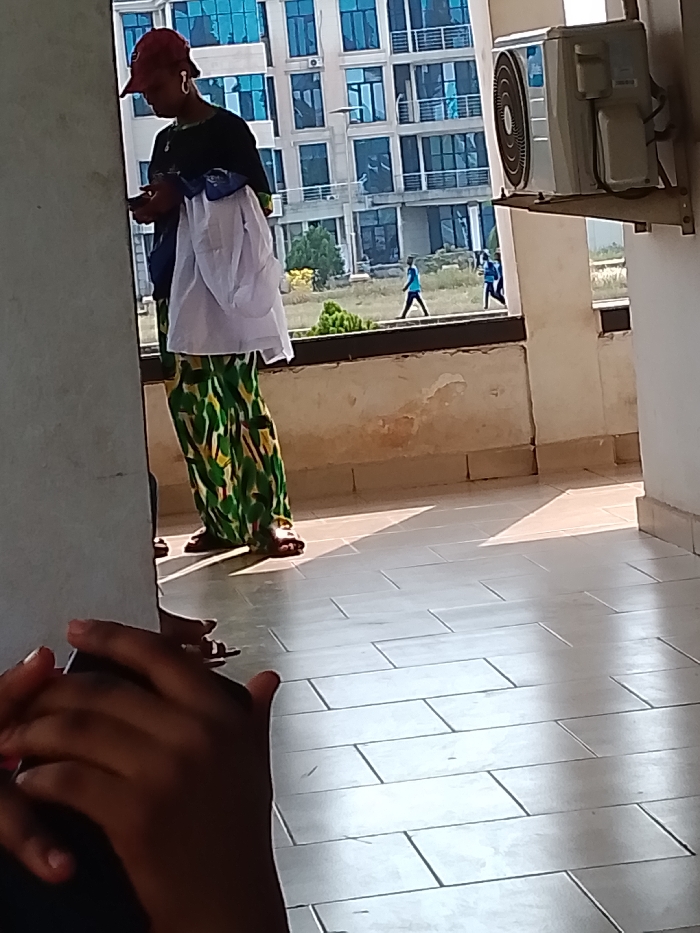 The image captures an outdoor scene in OGBOKO, Nigeria, under bright daylight. In the immediate foreground, dark-skinned hands are partially visible at the lower left. The ground is a light-colored tiled surface, reflecting significant sunlight, with rectangular tiles laid in a horizontal staggered pattern.
In the midground, a person stands predominantly on the left side, oriented slightly away from the camera. This individual wears a red baseball cap, a dark short-sleeved shirt, and patterned trousers or a long skirt featuring green, yellow, and black designs. White wireless earbuds are visible in their ears, and they are holding and looking at a mobile phone. A white garment is draped over their left arm. They wear dark sandals on their feet. Partially visible behind and to their left are another pair of feet wearing dark sandals, indicating a second, mostly obscured person.
To the right midground, an outdoor air conditioning unit is mounted high on a light-colored wall. Below this, a low, light-colored concrete barrier or railing separates the immediate area from the background, showing signs of wear and discoloration.
The background features an open area with sparse green shrubs and dry vegetation. Beyond this, a large, multi-story building with a modern design is visible, characterized by numerous windows with reflective blue glass. Two individuals are observed walking from left to right across the open ground between the barrier and the distant building. One person wears a light blue shirt and dark trousers, while the second, positioned further right, wears a dark blue shirt and dark trousers. Distinct shadows are cast on the tiled floor from an unseen light source to the left.