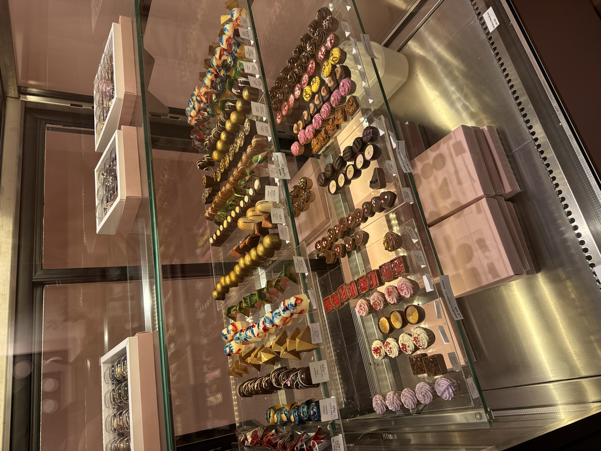 Here is a detailed description of the image:
**Scene**: The image shows a display case filled with assorted chocolates and candies. The chocolates are neatly arranged on shelves, showcasing a variety of colors, shapes, and designs. Boxes of chocolates are visible as well. The environment suggests an upscale confectionary or chocolate shop.
**Objects**:
* Assorted Chocolates: A wide variety of chocolates in different colors, shapes, and designs are arranged on the shelves.
* Boxes: Boxes of chocolates can be seen, particularly in the upper-right area of the case.
* Shelves: The shelves are made of glass.
* Price Tags: Several small price tags are placed next to the chocolates.
**Environment**:
* Location: The shop seems to be in Zagreb, Croatia, as some text contains the word "NARANČA" which is Croatian for "orange".
* Lighting: The lighting is bright, and internal to the case, highlighting the confectionery.
**Overall Impression**: The image conveys a sense of luxury and craftsmanship, typical of high-end chocolate shops.