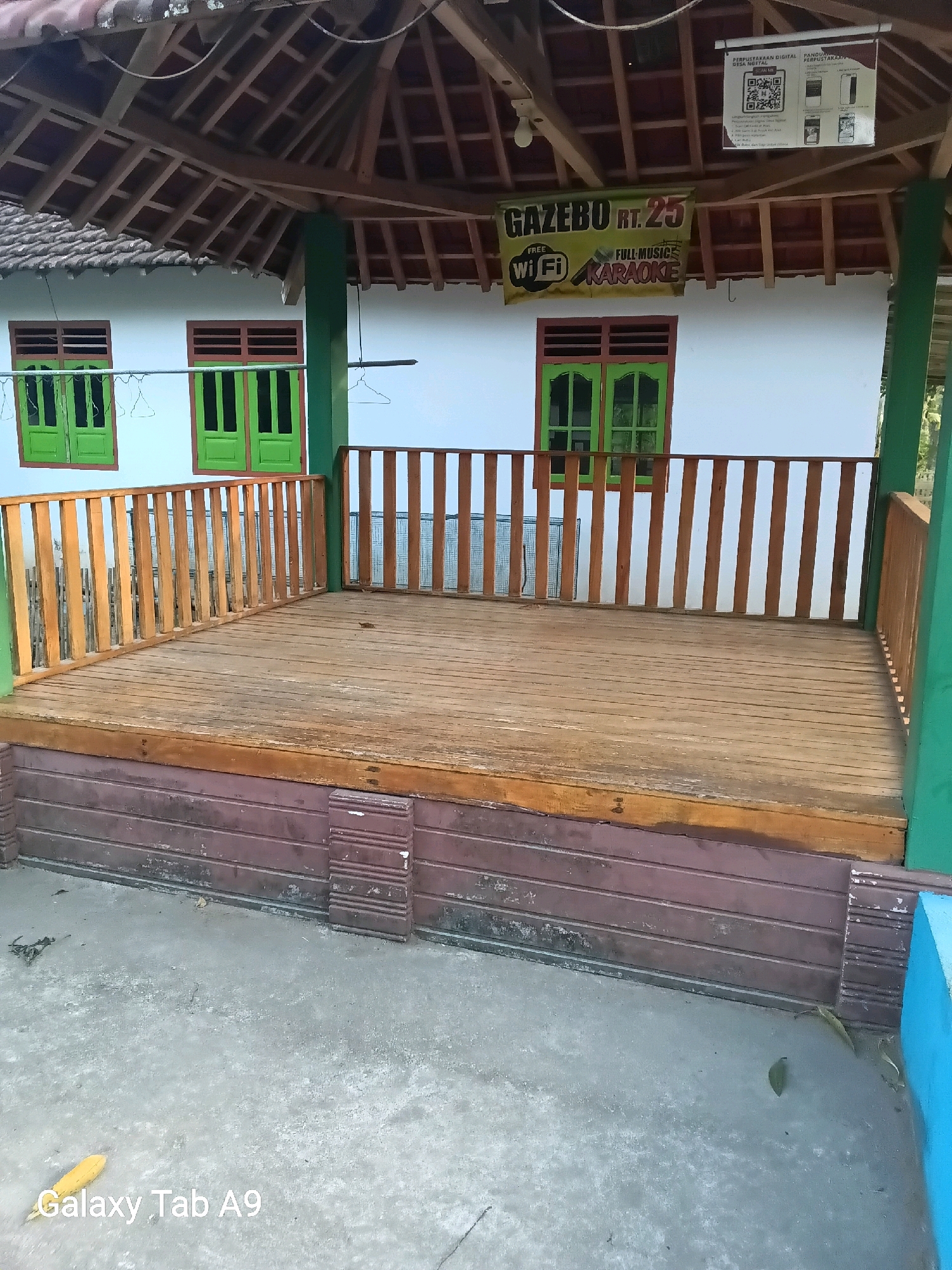 The image shows a wooden gazebo with a thatched roof, located in the city of Bendo, Indonesia. The gazebo has a raised wooden deck with wooden railings. On the white wall behind the gazebo, there are two green windows. A banner hangs from the roof, advertising "GAZEBO RT. 25 FREE WI FI FULL MUSIC KARAOKE." There is also a sign above the banner with information about a digital library. The ground in front of the gazebo is paved with concrete, and there is some debris on the ground. The overall scene suggests a public gathering space or a recreational area.