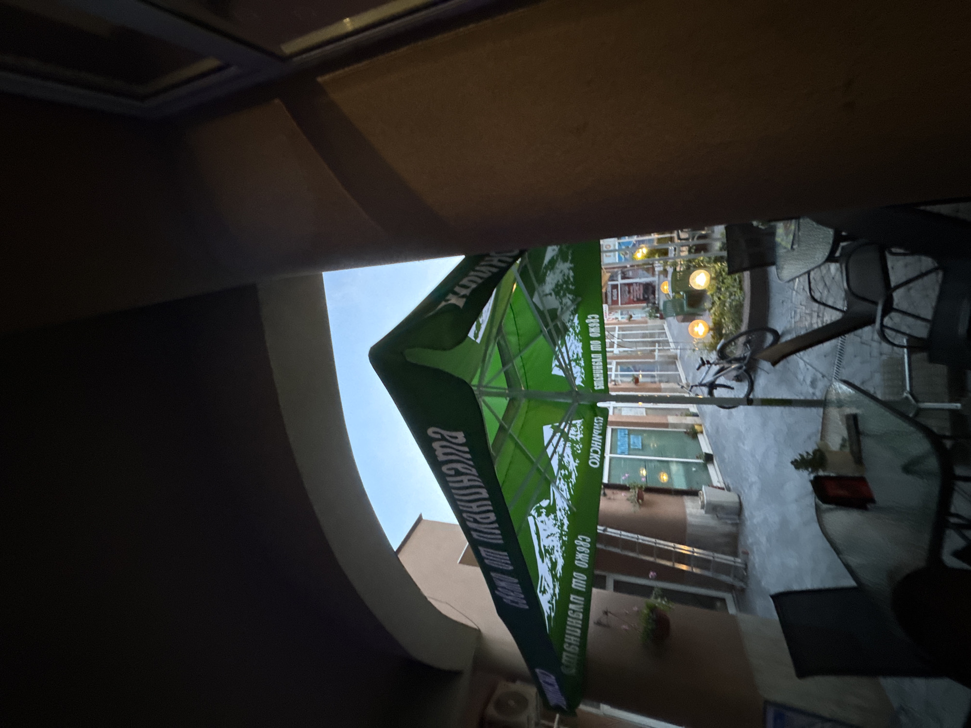 The image depicts an outdoor seating area of a establishment, likely a cafe or restaurant, situated in the city of Stamboliyski, Bulgaria. A large green umbrella, emblazoned with white text and a mountain landscape graphic, dominates the foreground. The umbrella casts a shadow over several tables and chairs arranged on a paved patio. In the background, a bicycle is parked near a set of illuminated globes. Buildings line the patio, suggesting an urban setting. The overall scene suggests a quiet, possibly evening, ambiance. The specific location within Stamboliyski is not explicitly identifiable from the visual information provided.